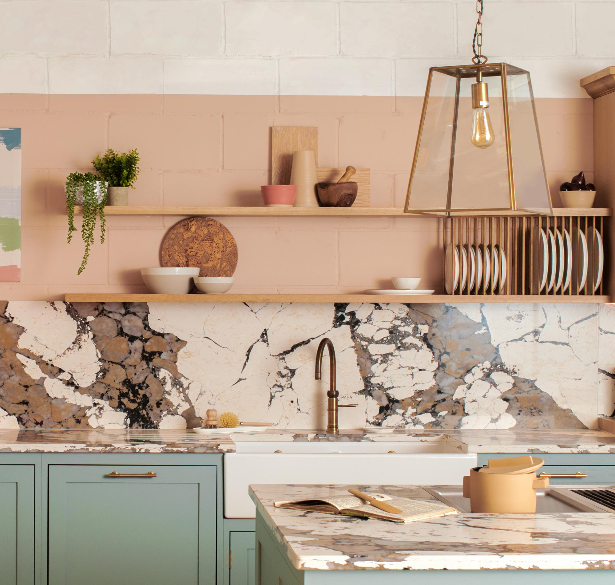 A modern kitchen with a marbled countertop and backsplash. Light blue lower cabinets contrast with a peach wall. Wooden shelves hold plants and dishes. A geometric pendant light hangs above a copper faucet and a beige kettle.