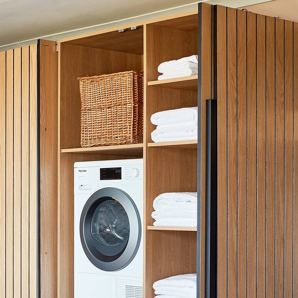 A modern laundry room features a washer and dryer set neatly into a wooden cabinet. Shelves beside the appliances hold neatly folded white towels and a wicker basket is placed on the top shelf.