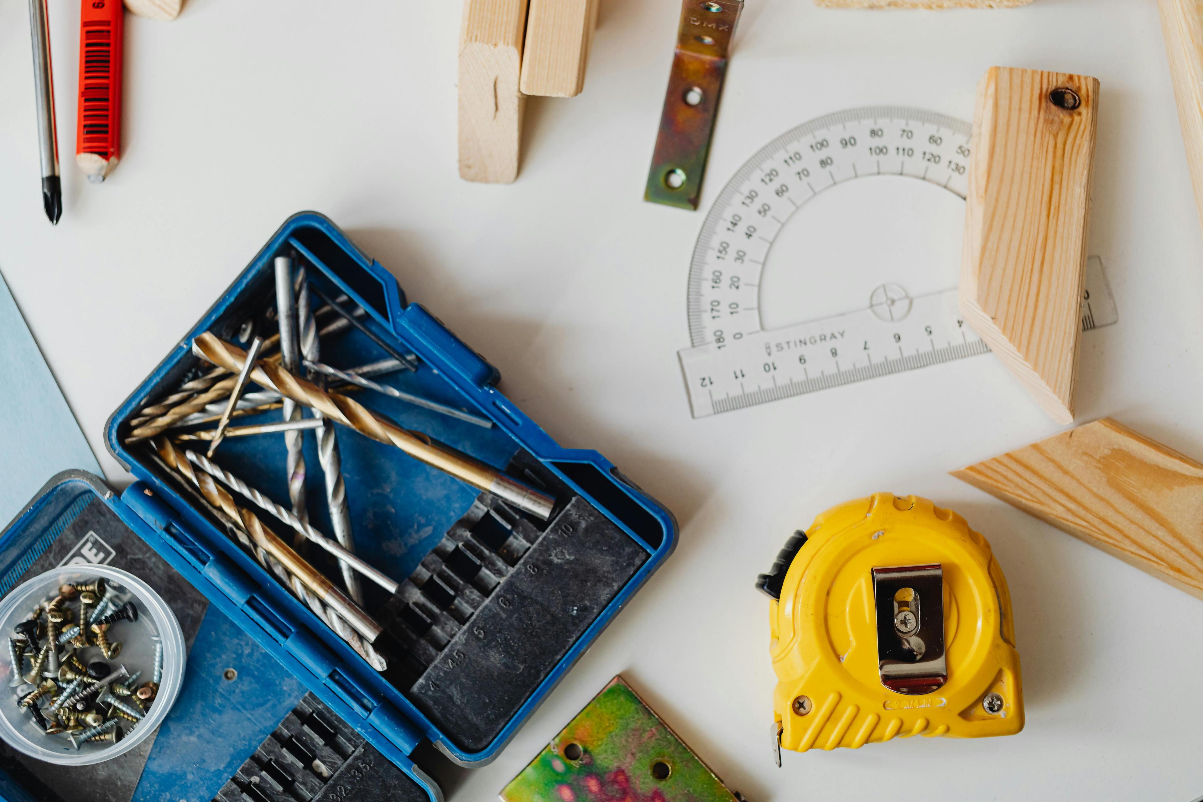 A flat lay overhead shot of carpentry tools, including an open blue case of drill bits, a small container of screws, a yellow tape measure, a clear plastic protractor, and pieces of cut wood on a white surface.
