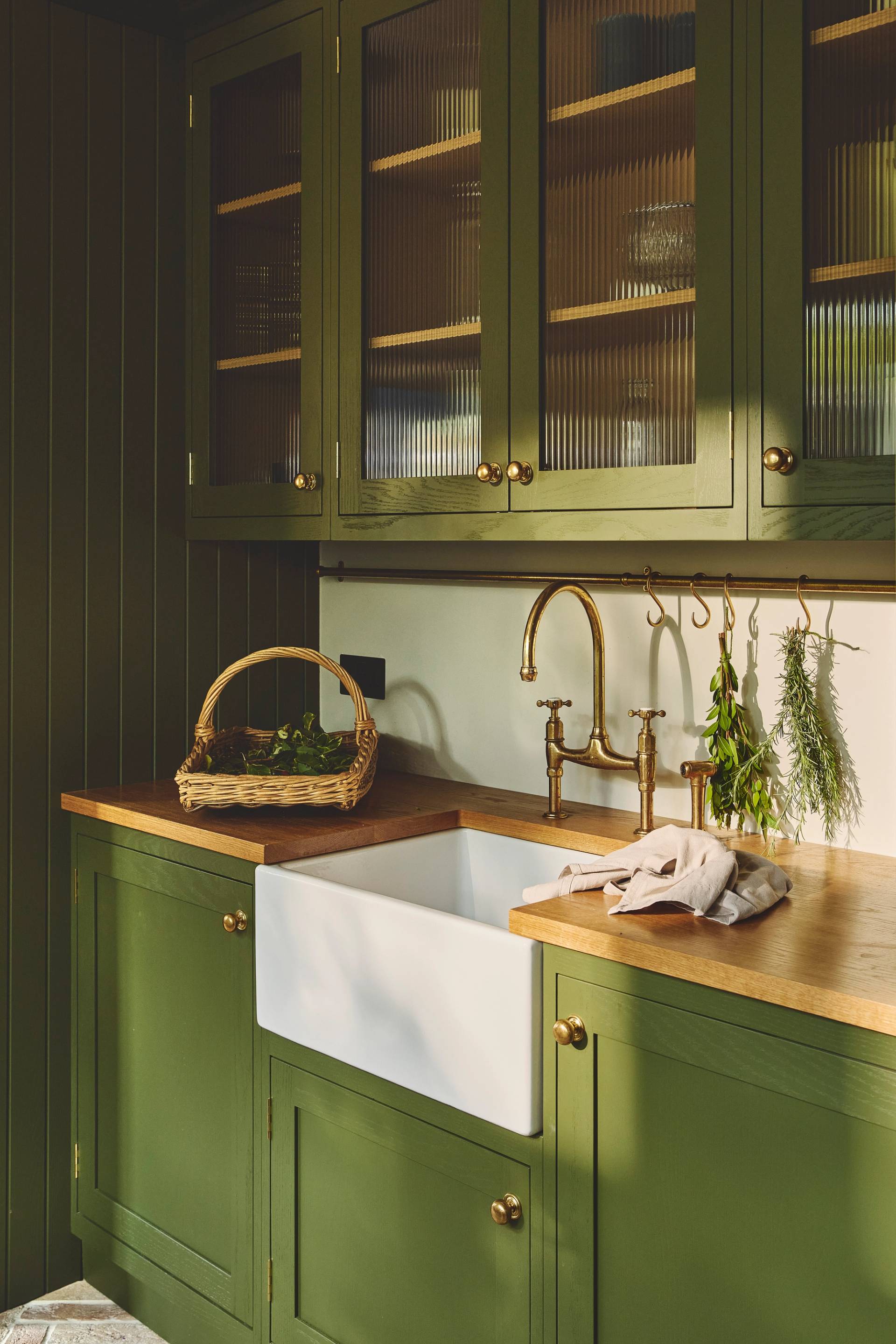 A cozy kitchen with olive green cabinets, brass hardware, a farmhouse sink, wooden countertops, a wicker basket, and fresh herbs hanging above the sink. Light filters in, highlighting the clean, rustic style.