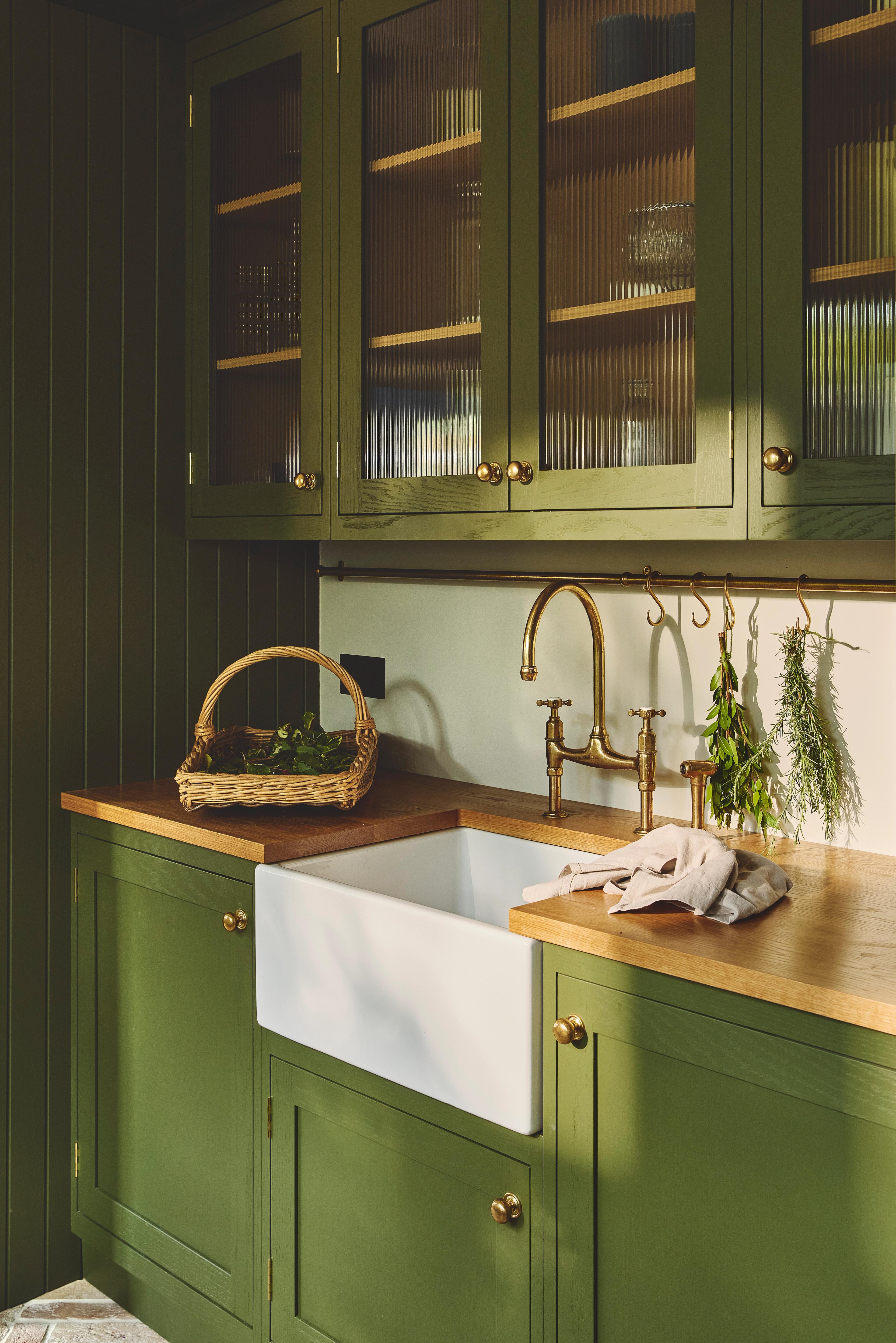 A cozy kitchen with olive green cabinets, brass hardware, a farmhouse sink, wooden countertops, a wicker basket, and fresh herbs hanging above the sink. Light filters in, highlighting the clean, rustic style.