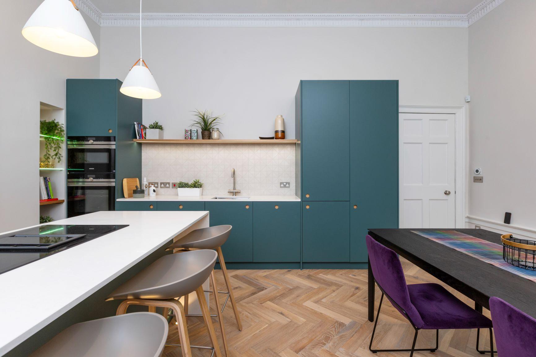 Modern kitchen with teal cabinets, white countertops, and wood-patterned flooring. Includes pendant lights, a sleek dining table with purple chairs, and open shelves with plants.