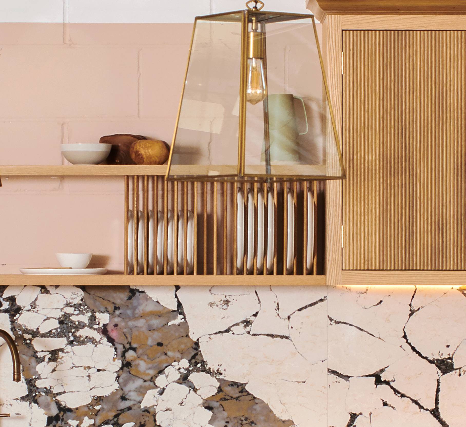 A modern kitchen with light pink walls features a wooden shelf holding white plates and bowls. Above, a geometric glass pendant light hangs. The countertop has a cracked stone pattern, creating a rustic look