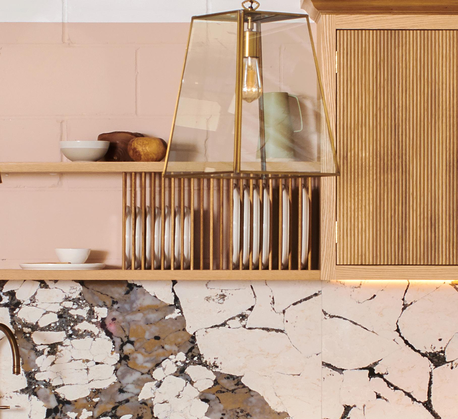 A modern kitchen with light pink walls features a wooden shelf holding white plates and bowls. Above, a geometric glass pendant light hangs. The countertop has a cracked stone pattern, creating a rustic look