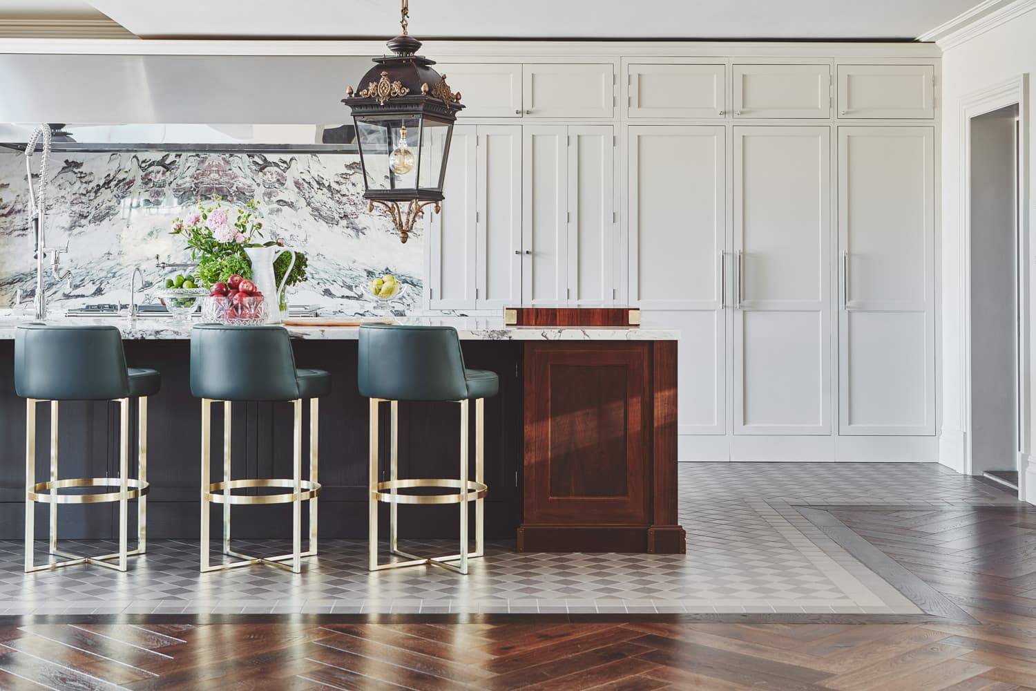 A modern kitchen with white cabinets, a large marble island, dark green barstools with gold bases, a wooden accent, and a black and gold pendant light above. The floor has a mix of tile and wood patterns.