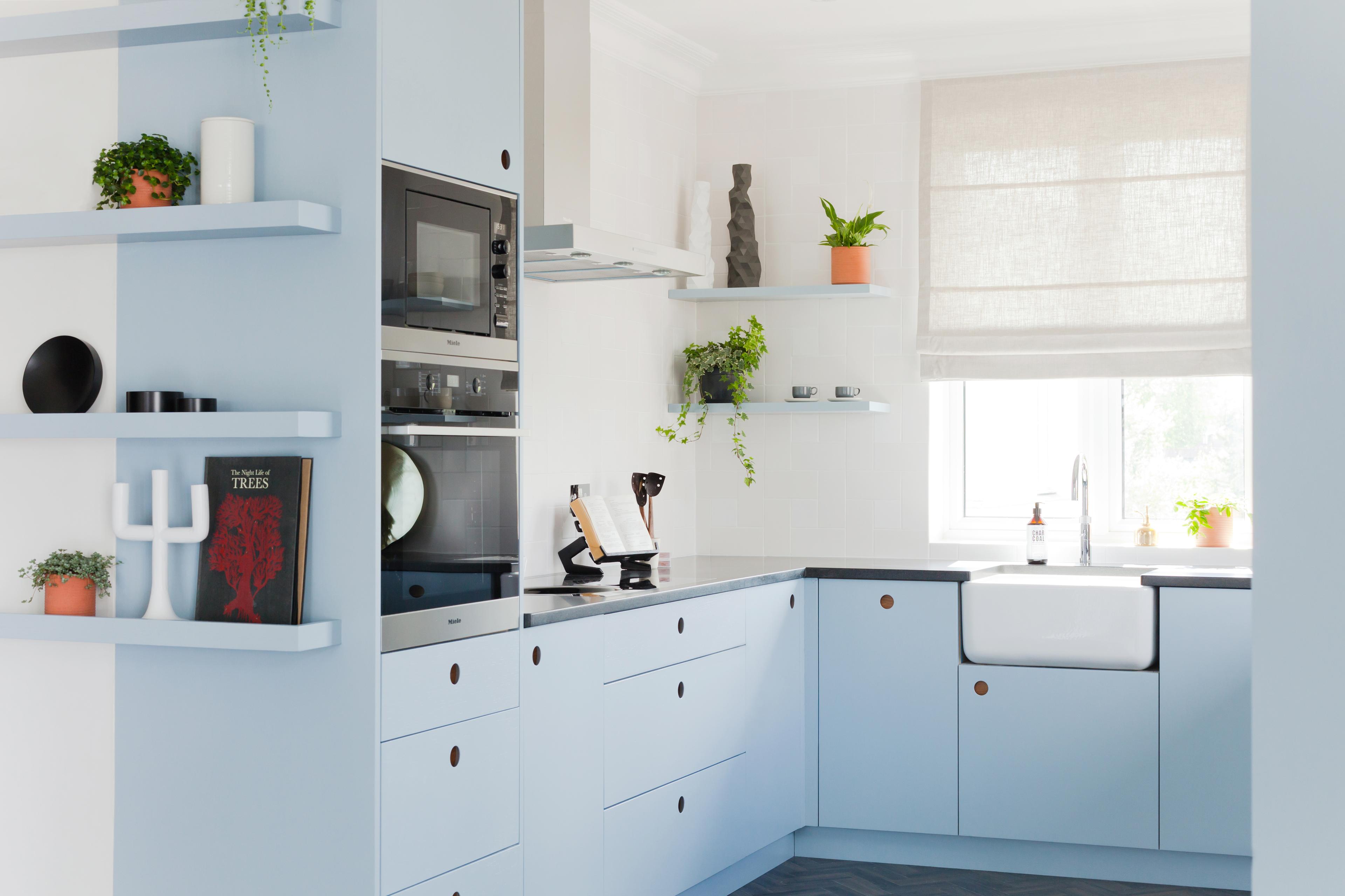 An angled view of a modern light blue kitchen featuring integrated stainless steel appliances, dark countertops, a white farmhouse sink beneath a bright window, and matching blue open shelving holding potted plants and decor.
