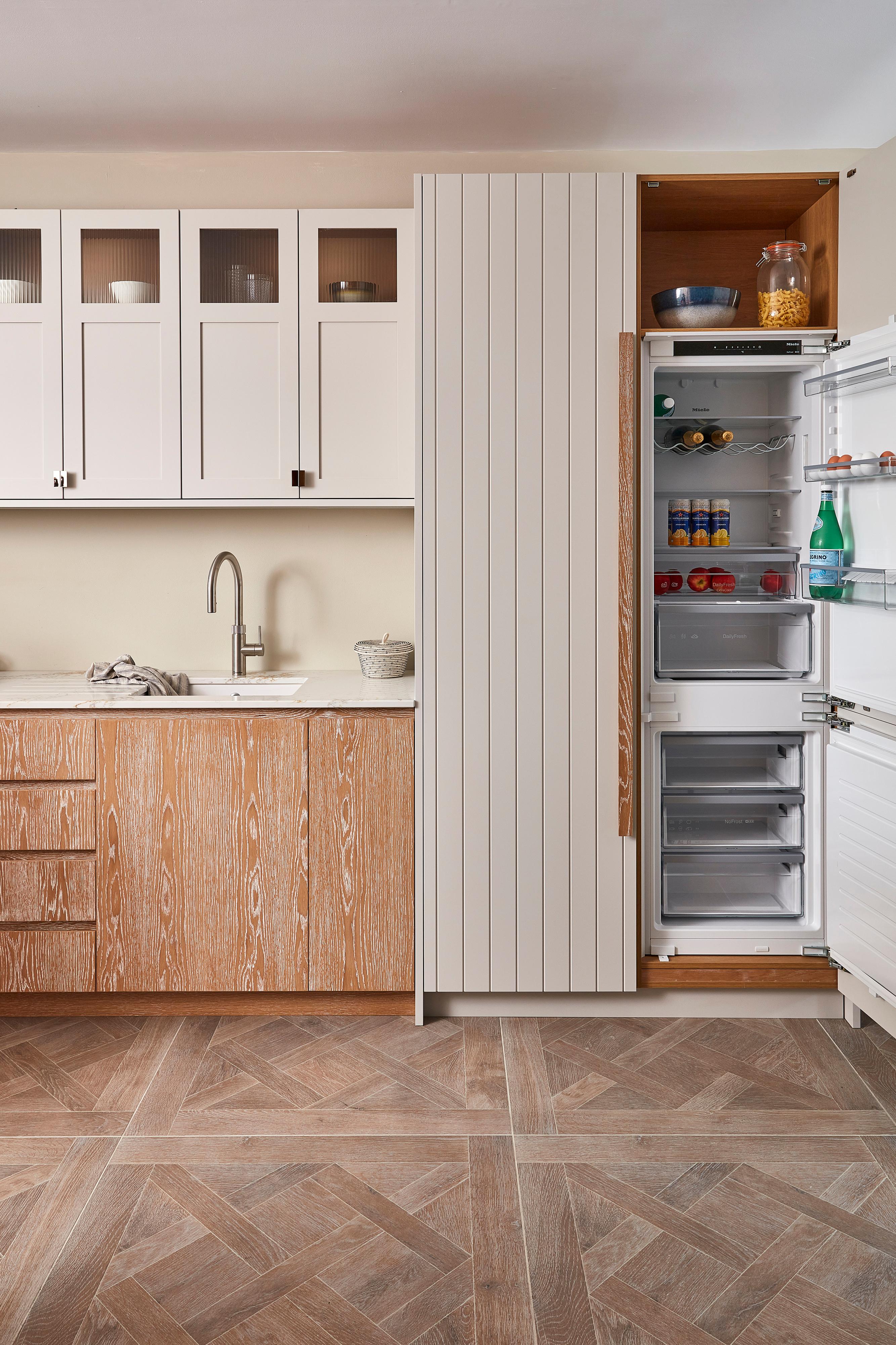A modern kitchen with white upper cabinets, wooden lower cabinets, and an open refrigerator stocked with various items. The floor features a geometric wood pattern, and a sink with a faucet is visible on the left