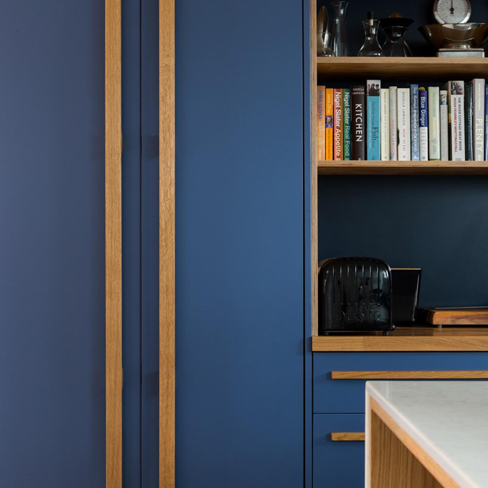 Modern kitchen with blue cabinets and wooden handles, an open shelf holding cookbooks and glassware, a black toaster, and a white countertop in the foreground.