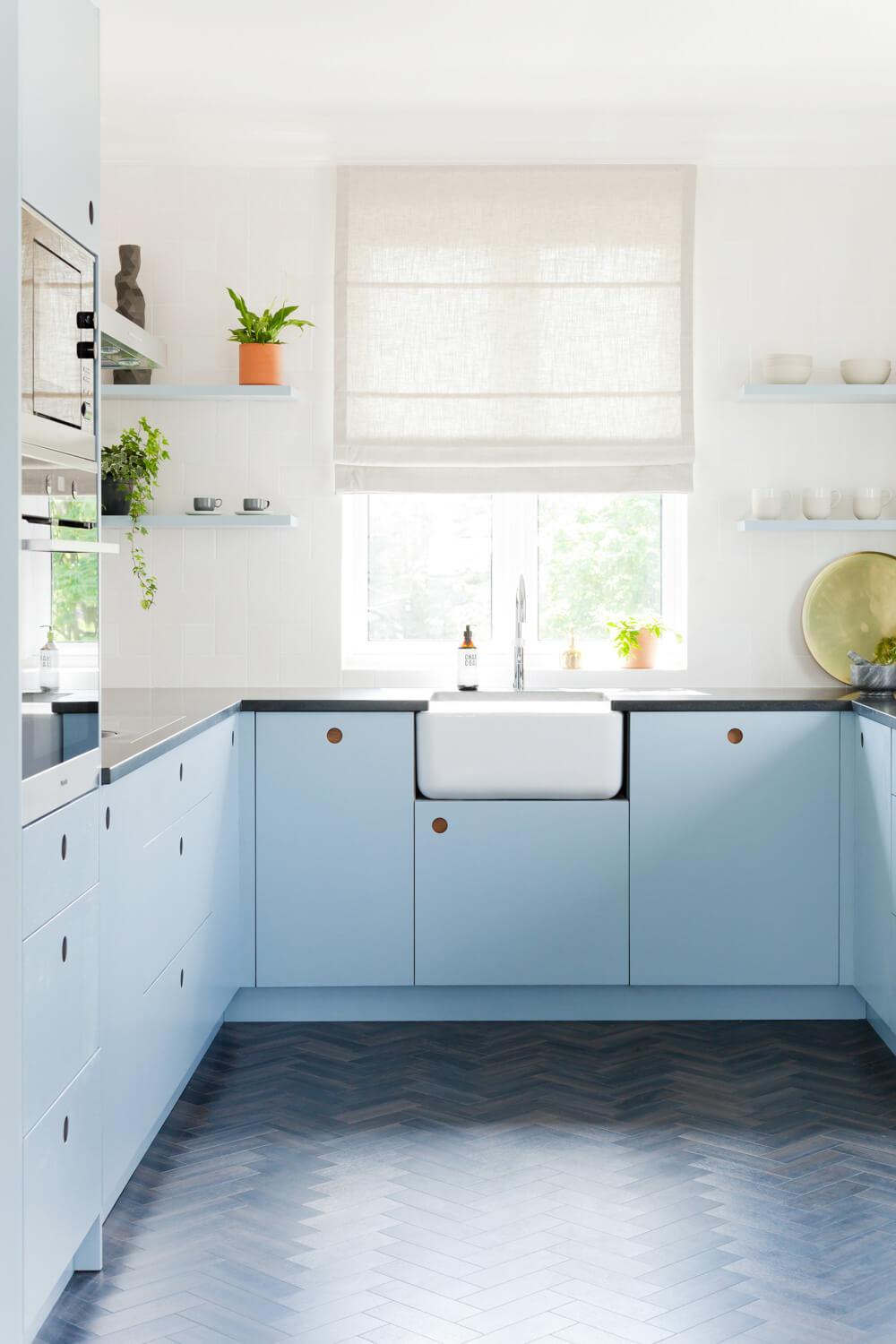A modern kitchen with light blue cabinets and black countertops. A white farmhouse sink is centered under a window with a beige Roman shade. Floating shelves hold potted plants and dishes. The floor features a dark blue herringbone pattern. Natural light fills the space.