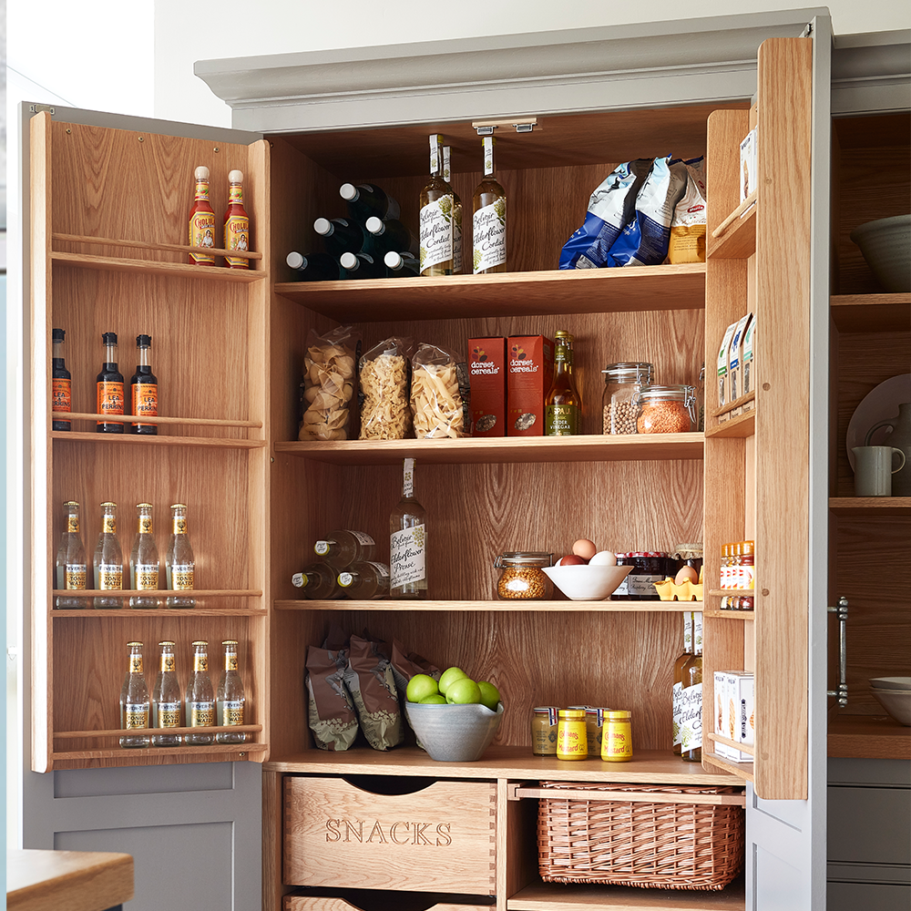 A neatly organized pantry with open wooden doors displays bottles, jars, pasta, chips, snacks, fruit, and drinks on shelves, with wooden crates labeled “SNACKS” and a wicker basket at the bottom.