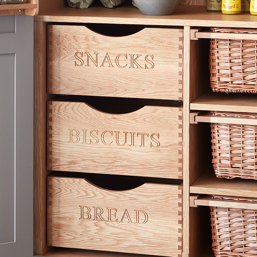Three wooden drawers labeled SNACKS, BISCUITS, and BREAD are built into a shelving unit, with wicker baskets on shelves beside them.