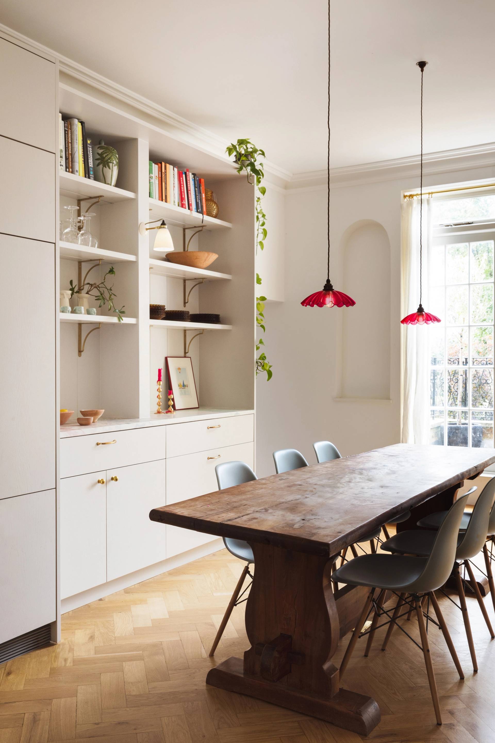 A dining room with a wooden table and eight blue chairs. Built-in white shelves display books and decor. Two hanging red lamps provide light. A window with closed curtains lets in daylight, and a plant vine drapes from the shelf.