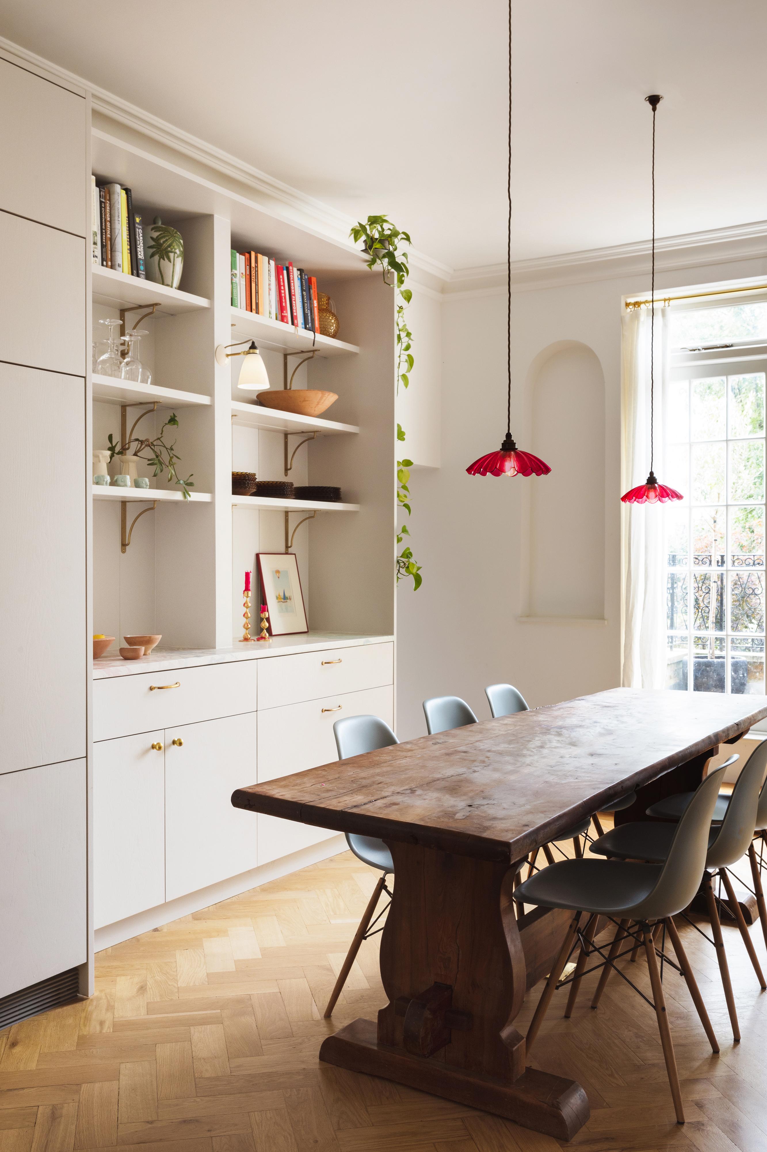 A dining room with a wooden table and eight blue chairs. Built-in white shelves display books and decor. Two hanging red lamps provide light. A window with closed curtains lets in daylight, and a plant vine drapes from the shelf.