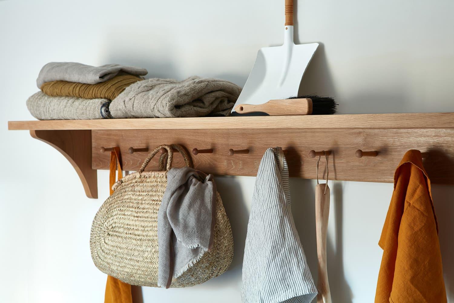A wooden wall shelf with several hooks holds folded blankets and clothes. A dustpan and brush are on top, and a woven straw bag, scarf, and orange clothing hang from the hooks.