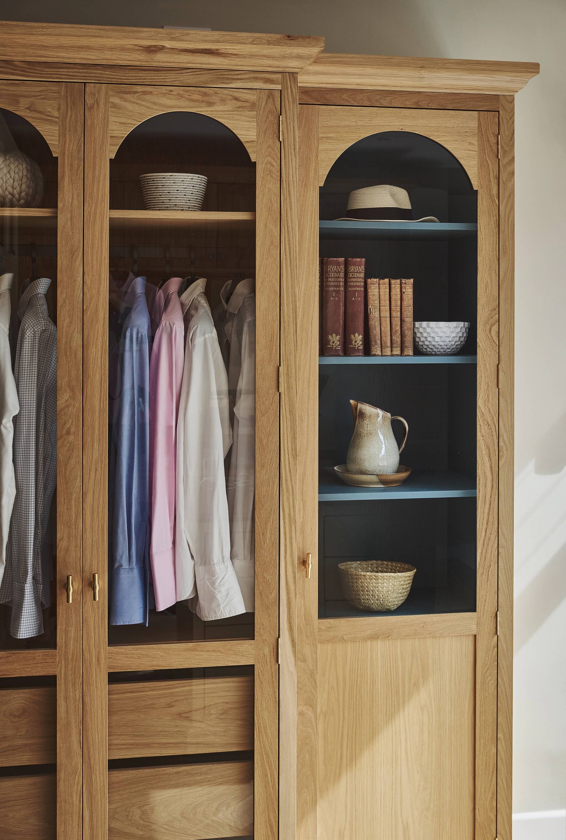 An oak wardrobe with glass doors displays neatly hung shirts. Adjacent open shelves hold books, a hat, a jug, a basket, and a wicker box, creating an organized and cozy storage space.
