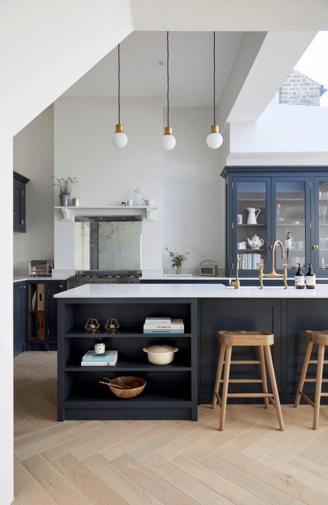 Modern kitchen with dark cabinets, a large island, and three wooden stools. Pendant lights hang above the island, and a skylight brightens the space. Open shelves display kitchenware, and a radiator is visible on the right wall.