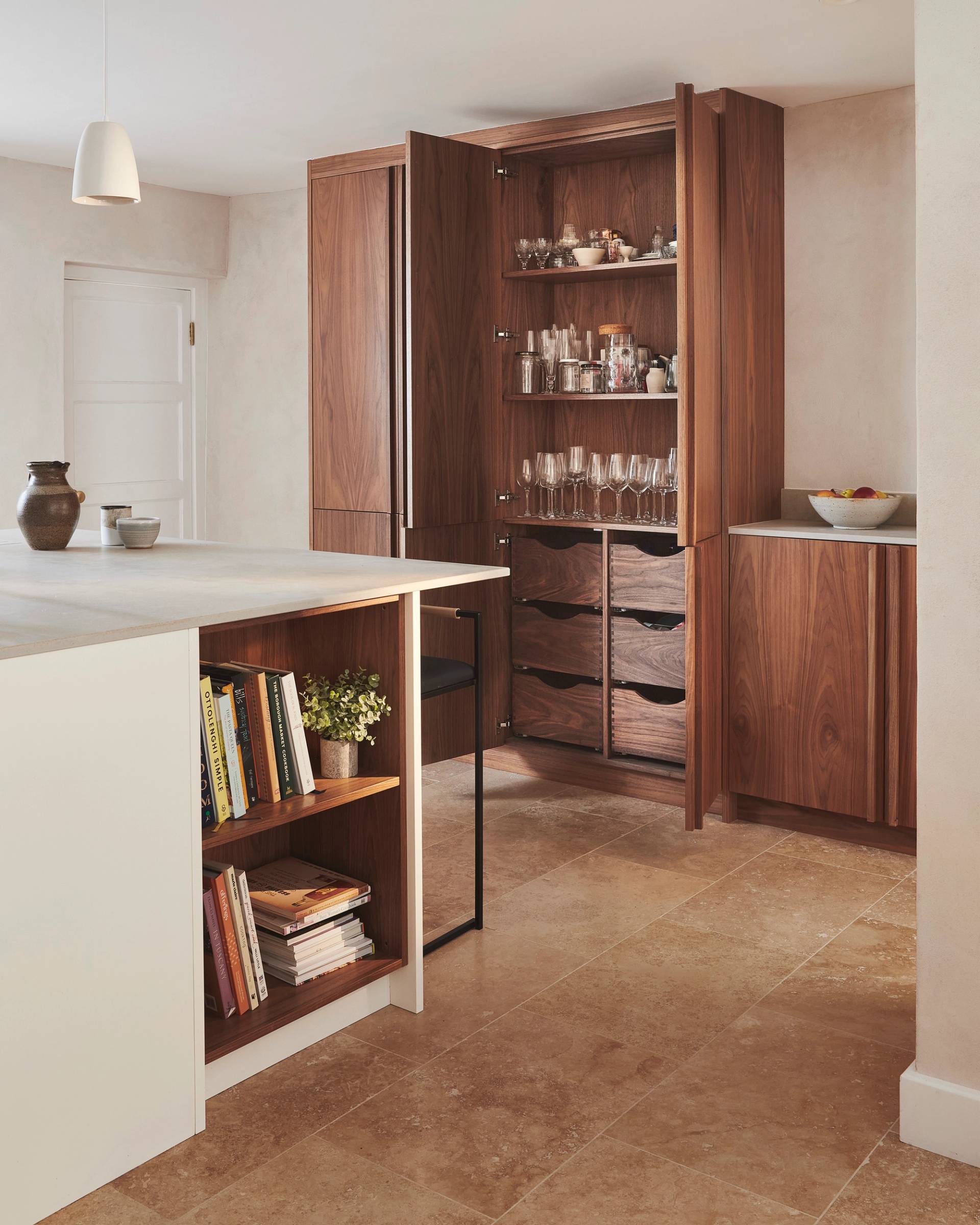 Modern kitchen with a minimalist design. Features wooden cabinets, a large island with a light countertop, and a few decorative items. Open shelving on island holds books. Beige stone tile flooring enhances the warm, natural aesthetic.