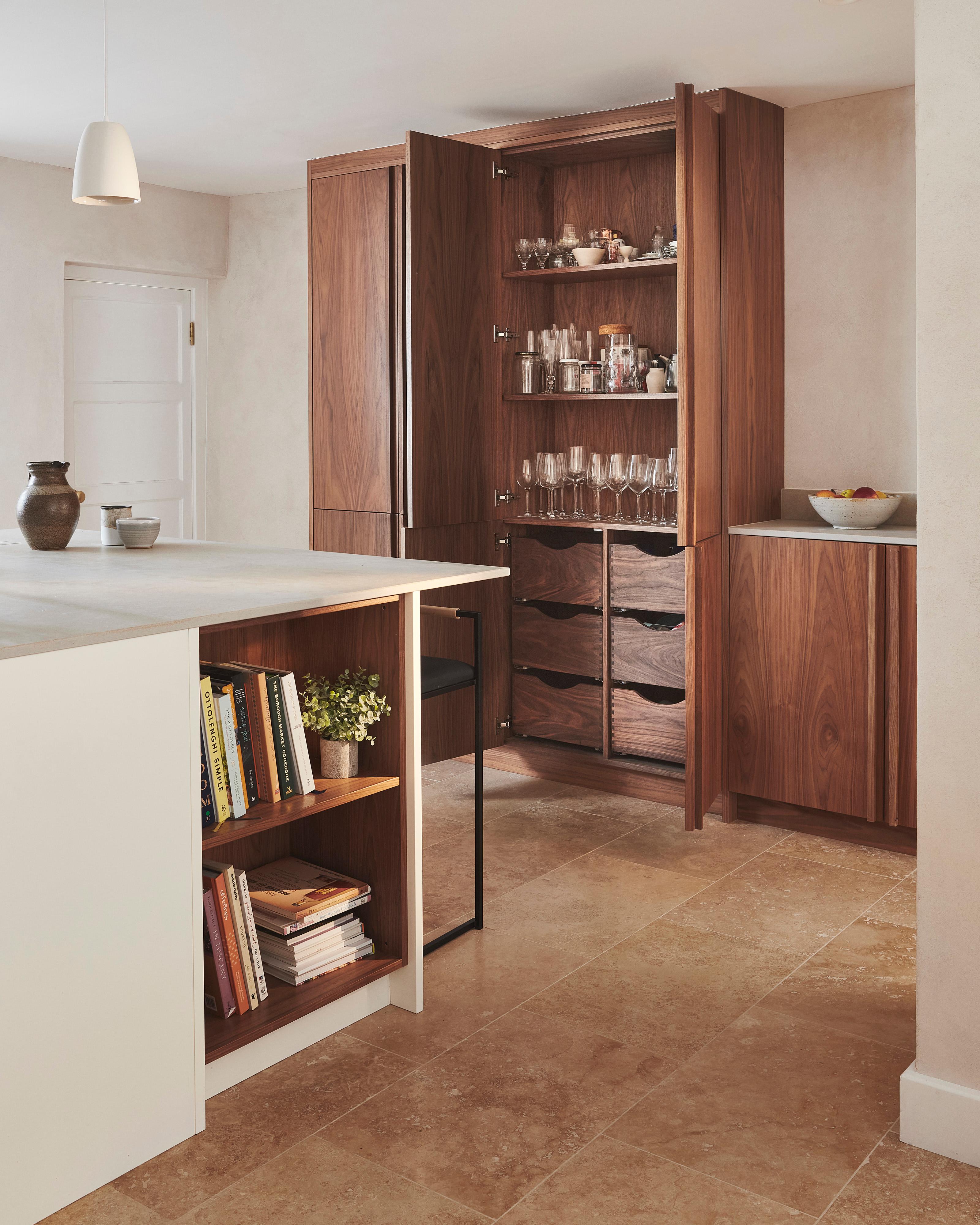 Modern kitchen with a minimalist design. Features wooden cabinets, a large island with a light countertop, and a few decorative items. Open shelving on island holds books. Beige stone tile flooring enhances the warm, natural aesthetic.