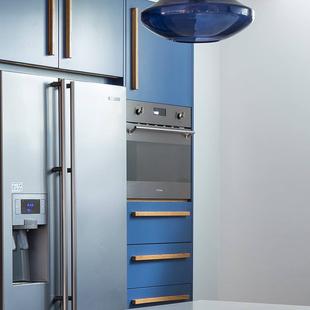 A modern kitchen with blue cabinets, a stainless steel refrigerator with a water dispenser, a built-in oven, and a blue pendant light overhead.