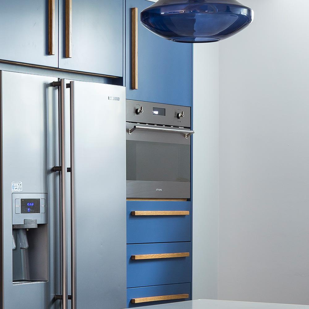 A modern kitchen with blue cabinets, a stainless steel refrigerator with a water dispenser, a built-in oven, and a blue pendant light overhead.