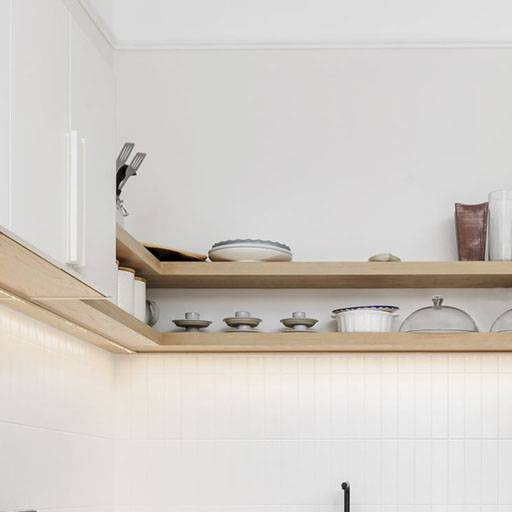 Two wooden kitchen shelves display stacked plates, bowls, cups, utensils, and covered dishes against a white tiled wall and cabinetry in a minimalist, modern kitchen setting.