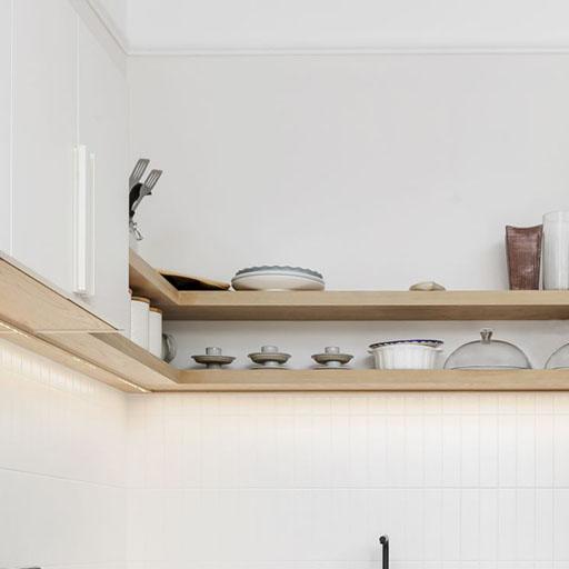 Two wooden kitchen shelves display stacked plates, bowls, cups, utensils, and covered dishes against a white tiled wall and cabinetry in a minimalist, modern kitchen setting.