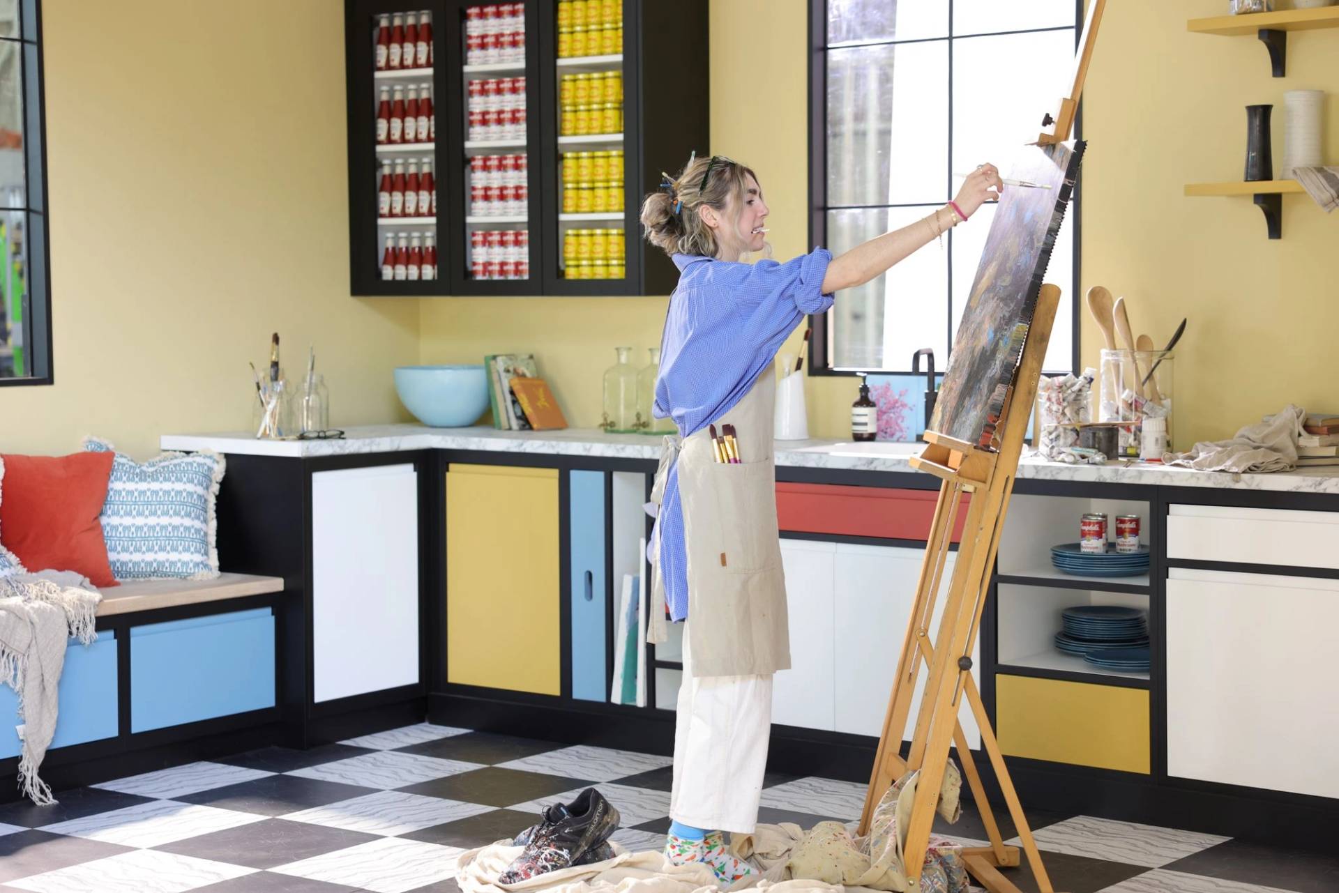 An artist in a blue shirt and apron paints on a wooden easel in a bright, modern studio. The room features Mondrian-inspired color-blocked cabinets, checkered floors, and shelves stocked with colorful soup cans.