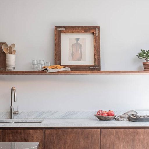 A modern kitchen with a marble countertop, wooden cabinets, a sink, and an open wooden shelf holding glasses, a potted plant, kitchen utensils, and framed art. A bowl of pomegranates sits on the counter.
