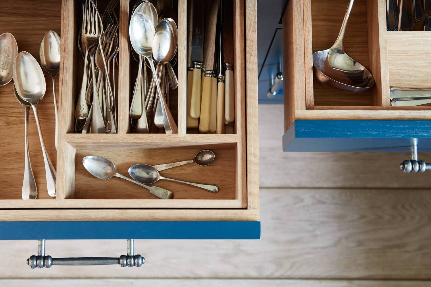 Open kitchen drawers reveal neatly organized cutlery, including spoons, forks, and knives in wooden compartments. Silverware is prominently displayed, showcasing different sizes and styles, with modern blue drawer fronts and silver handles.
