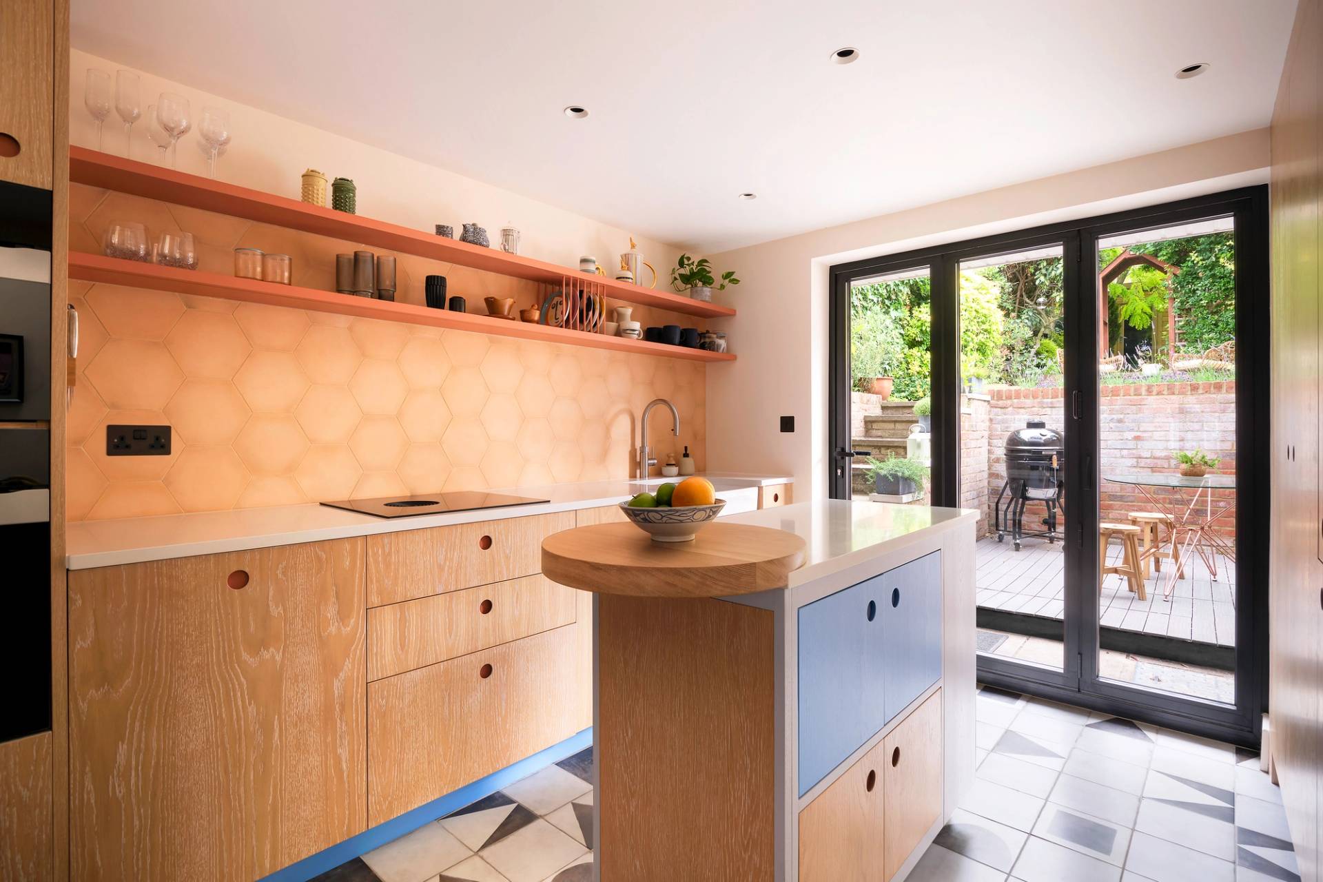 A modern kitchen with wooden cabinets and a circular island topped with a fruit bowl. Open shelving displays glassware and ceramics. Hexagonal tiles form the backsplash. Sliding glass doors lead to a patio with outdoor furniture and greenery.