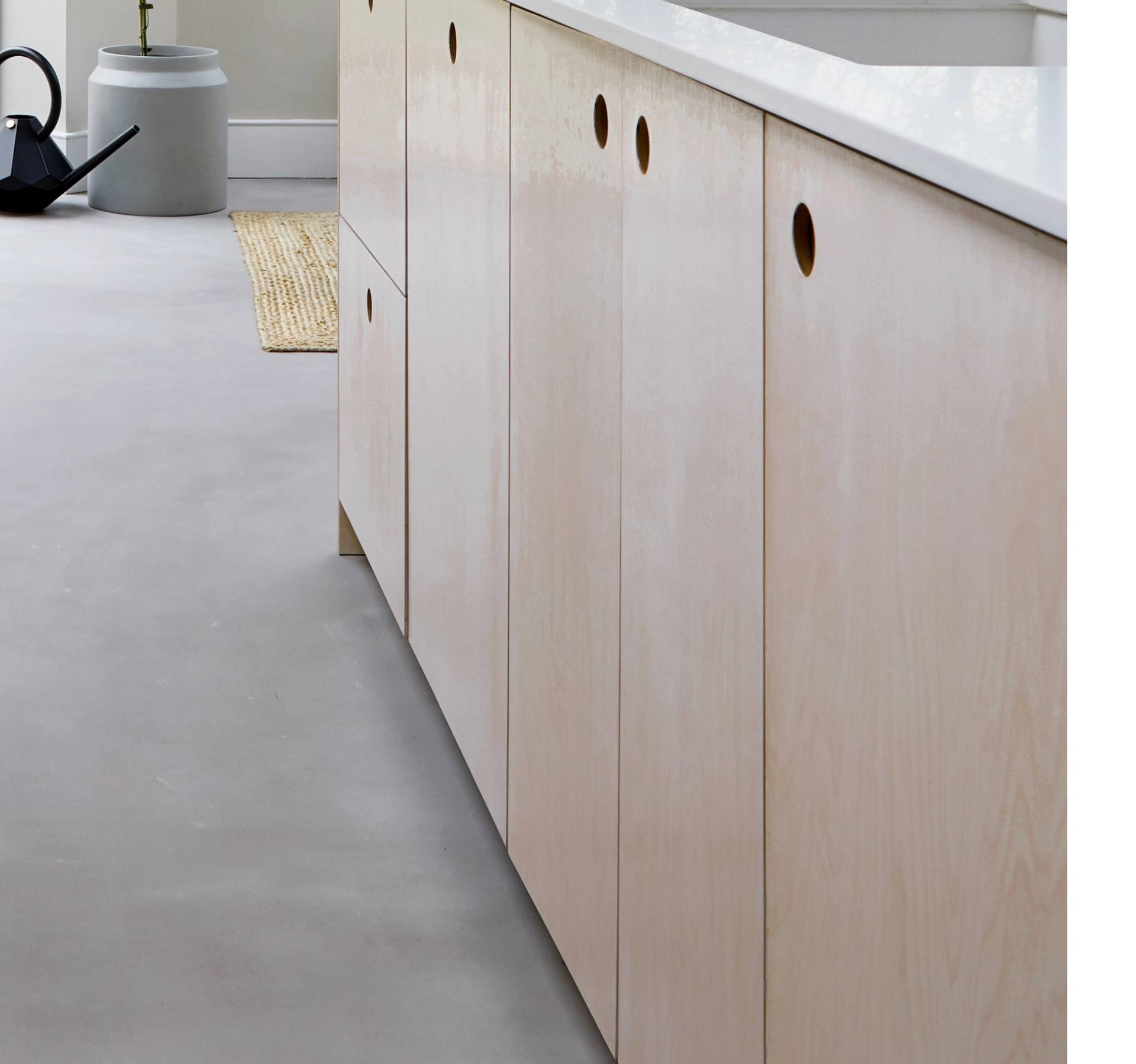 Light wood kitchen cabinets with round cut-out handles are shown against a light grey floor. A black watering can, a woven mat, and a grey vase with a plant appear in the background near a white wall.