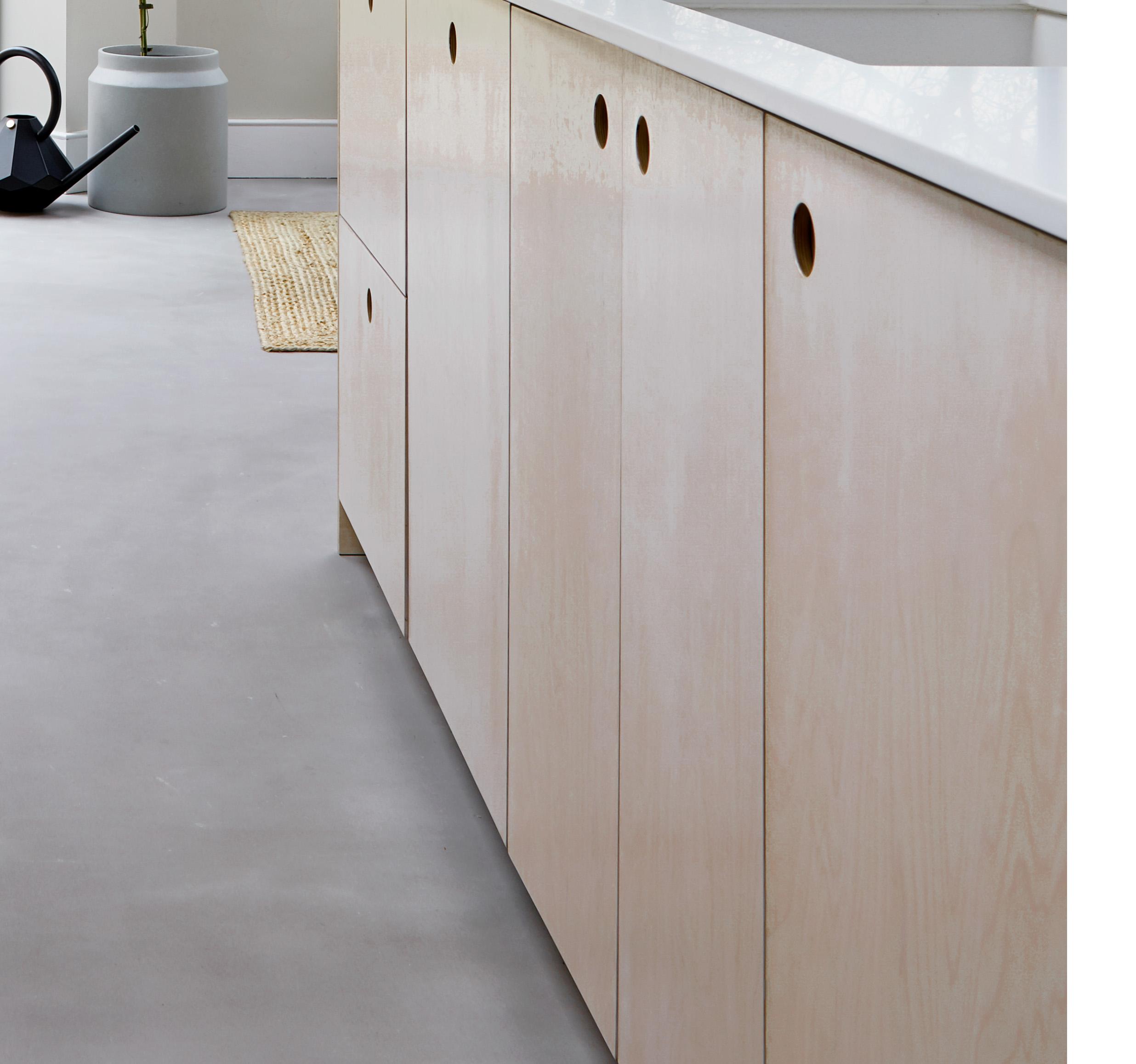 Light wood kitchen cabinets with round cut-out handles are shown against a light grey floor. A black watering can, a woven mat, and a grey vase with a plant appear in the background near a white wall.