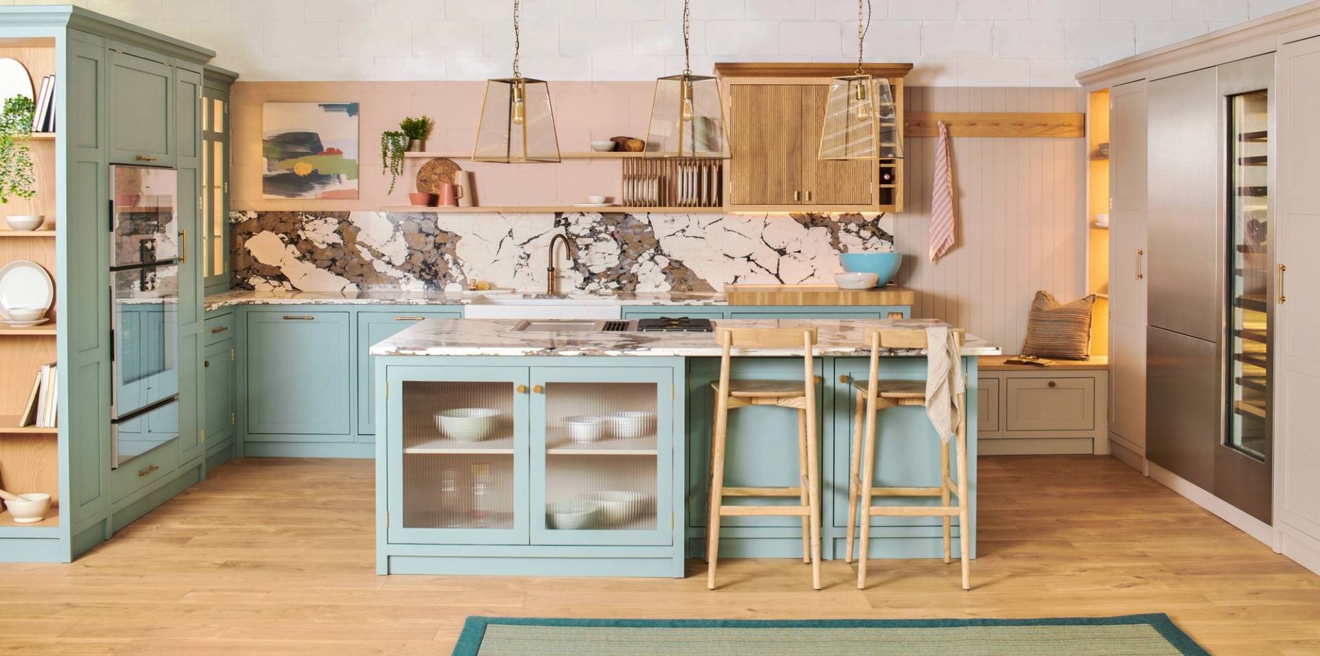 A cozy kitchen with pastel blue cabinets, a marble backsplash and countertop, wooden stools at an island, hanging pendant lights, and open shelving decorated with dishes and plants.