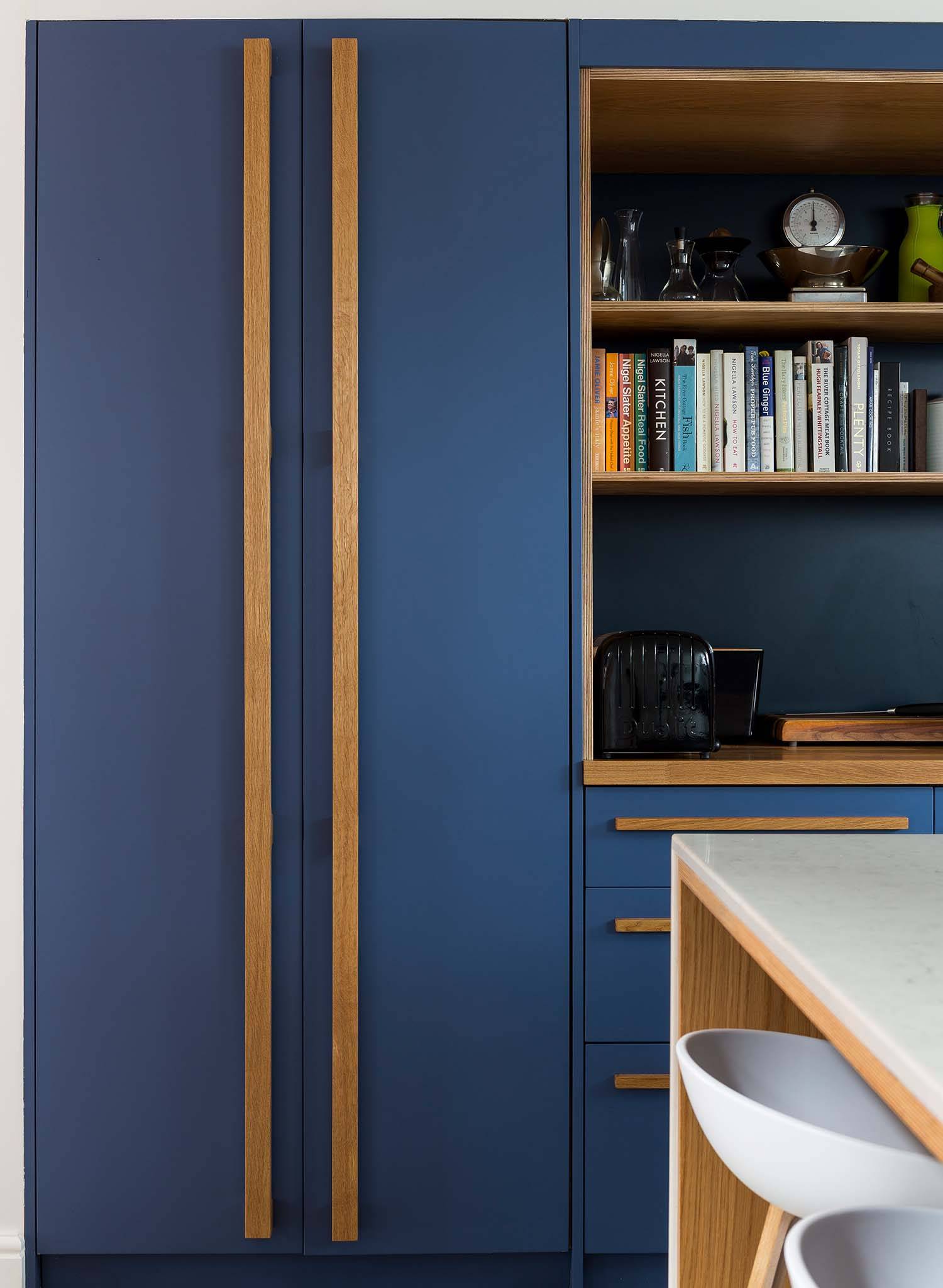 A modern kitchen with matte blue cabinets, long wooden handles, open shelves with books and kitchenware, a black toaster, and a white countertop with barstools.