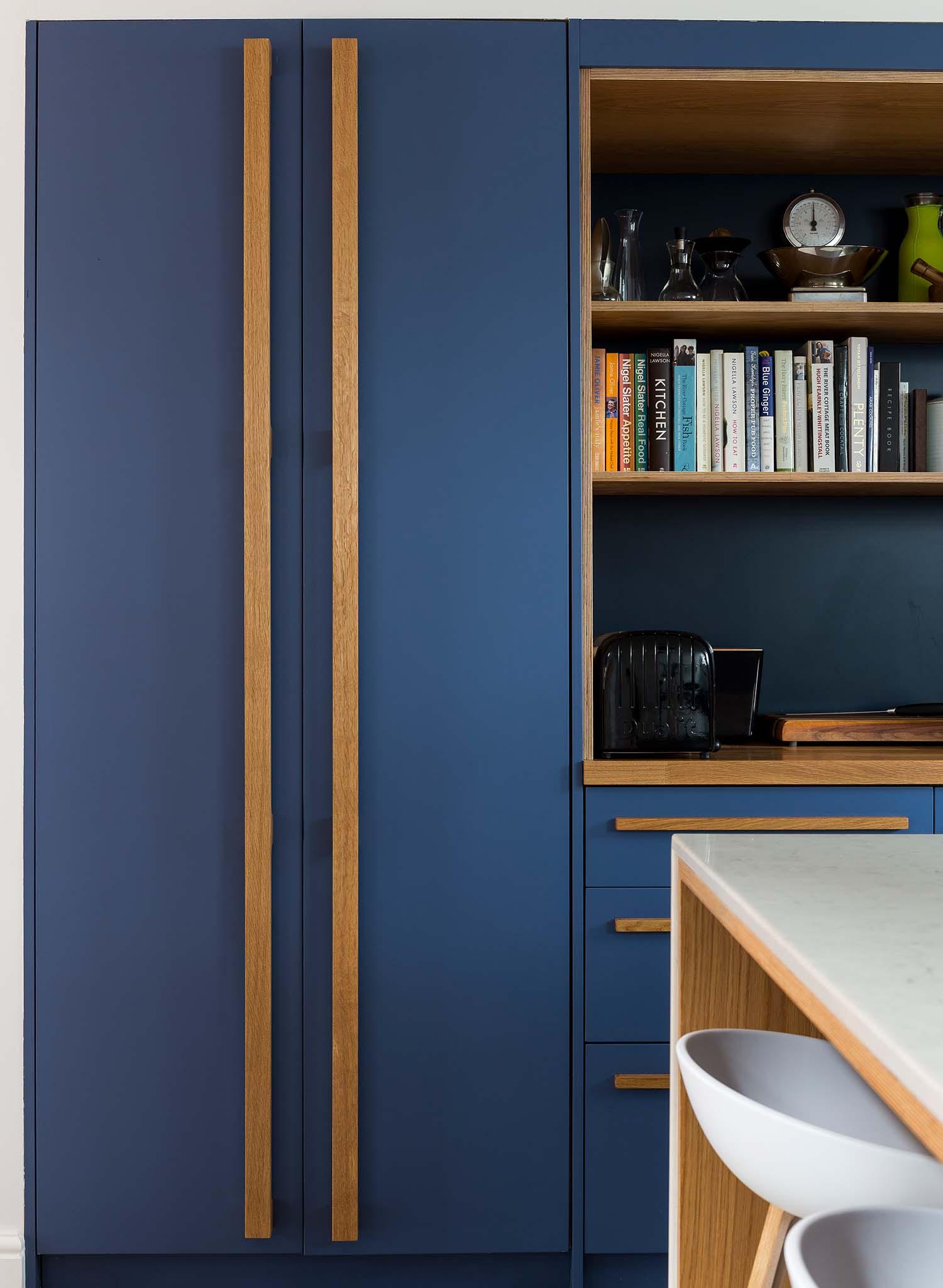 A modern kitchen with matte blue cabinets, long wooden handles, open shelves with books and kitchenware, a black toaster, and a white countertop with barstools.