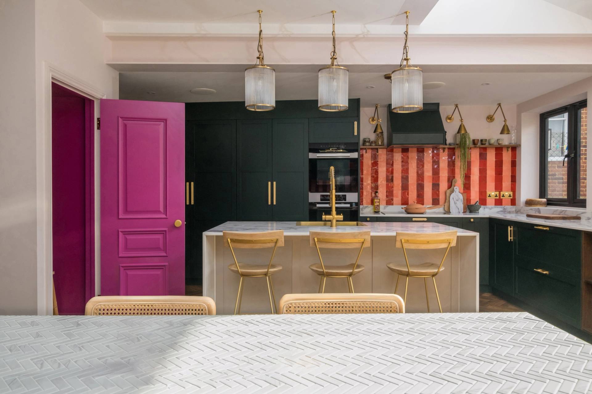 A modern kitchen with dark green cabinets, a central island with gold bar stools, and a colorful red and orange backsplash. Three glass pendant lights hang overhead, and two candles on wooden holders sit on a patterned white tabletop. A vibrant magenta door is open on the left.