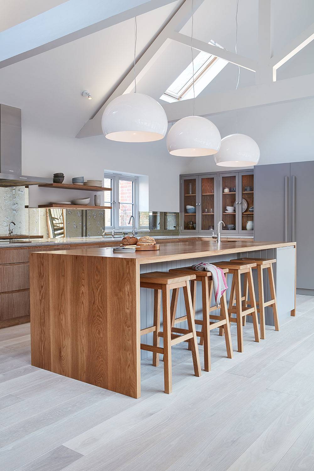 Modern kitchen with a wooden island, bar stools, and white countertops. Three large white pendant lights hang from the ceiling. Light wood flooring complements the minimalistic design, and a skylight brightens the space.