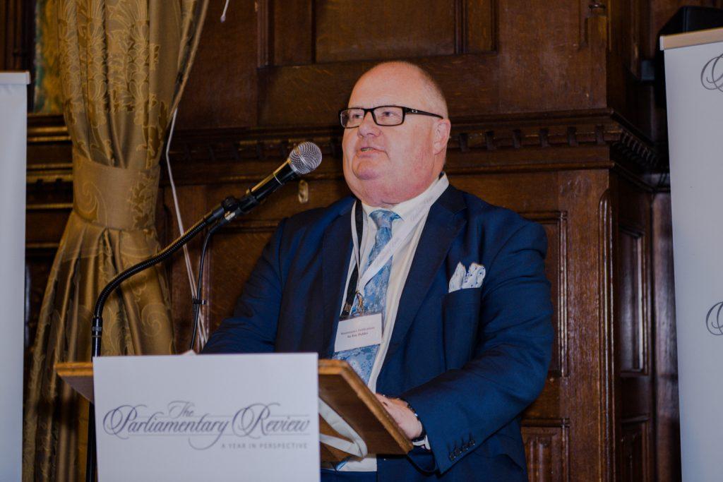 A man wearing glasses and a blue suit speaks at a wooden podium featuring "The Parliamentary Review" sign.