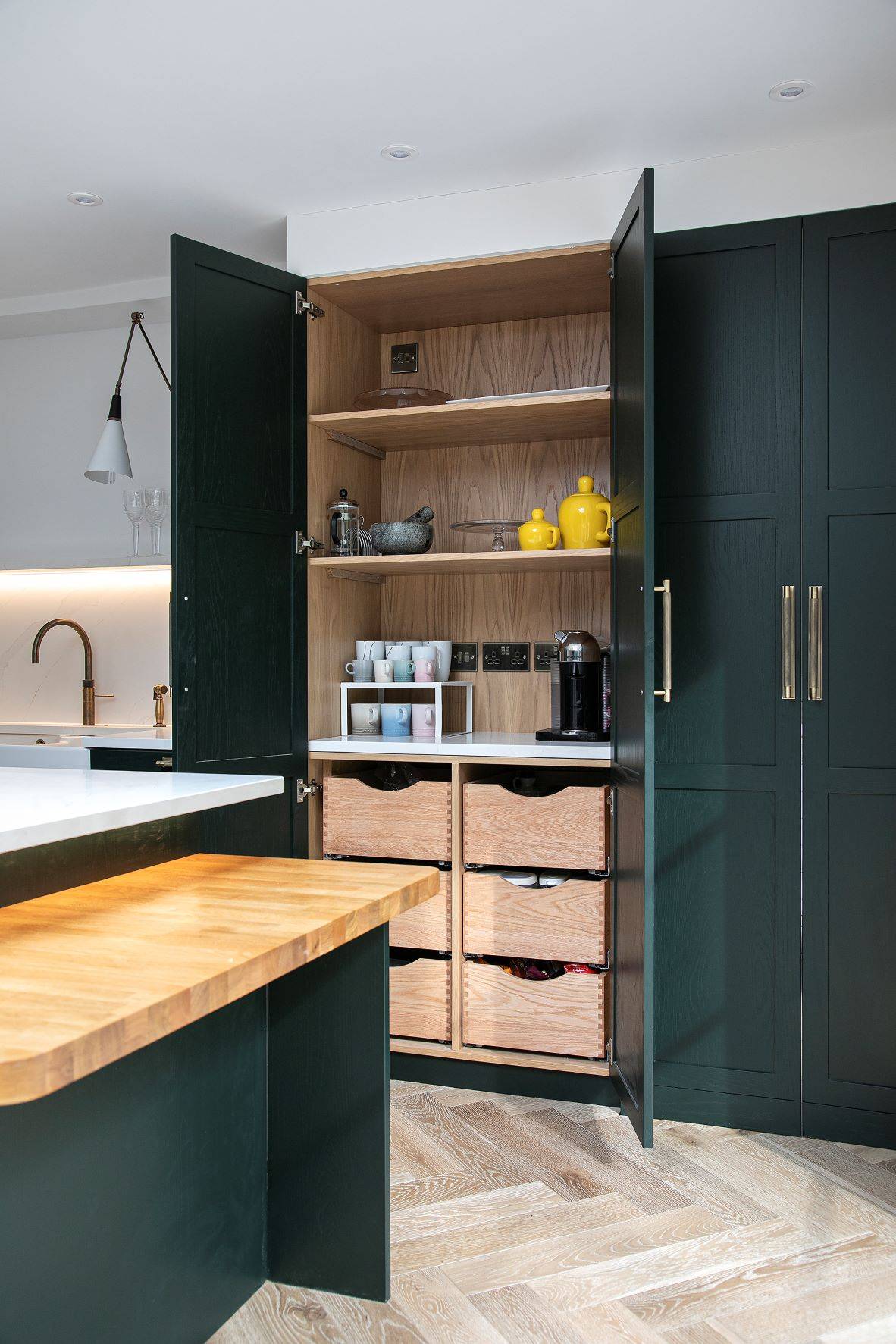 Modern kitchen with dark green cabinets, open pantry showing shelves with mugs, yellow teapots, a coffee machine, and four wooden drawers. Theres a wooden counter and herringbone wood flooring