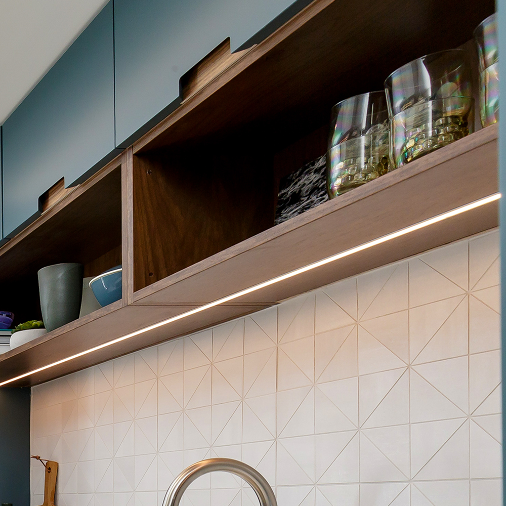 A modern kitchen with open wooden shelves holding glassware and bowls. Below, a textured white tile backsplash and under-cabinet LED lighting are visible above a silver faucet.