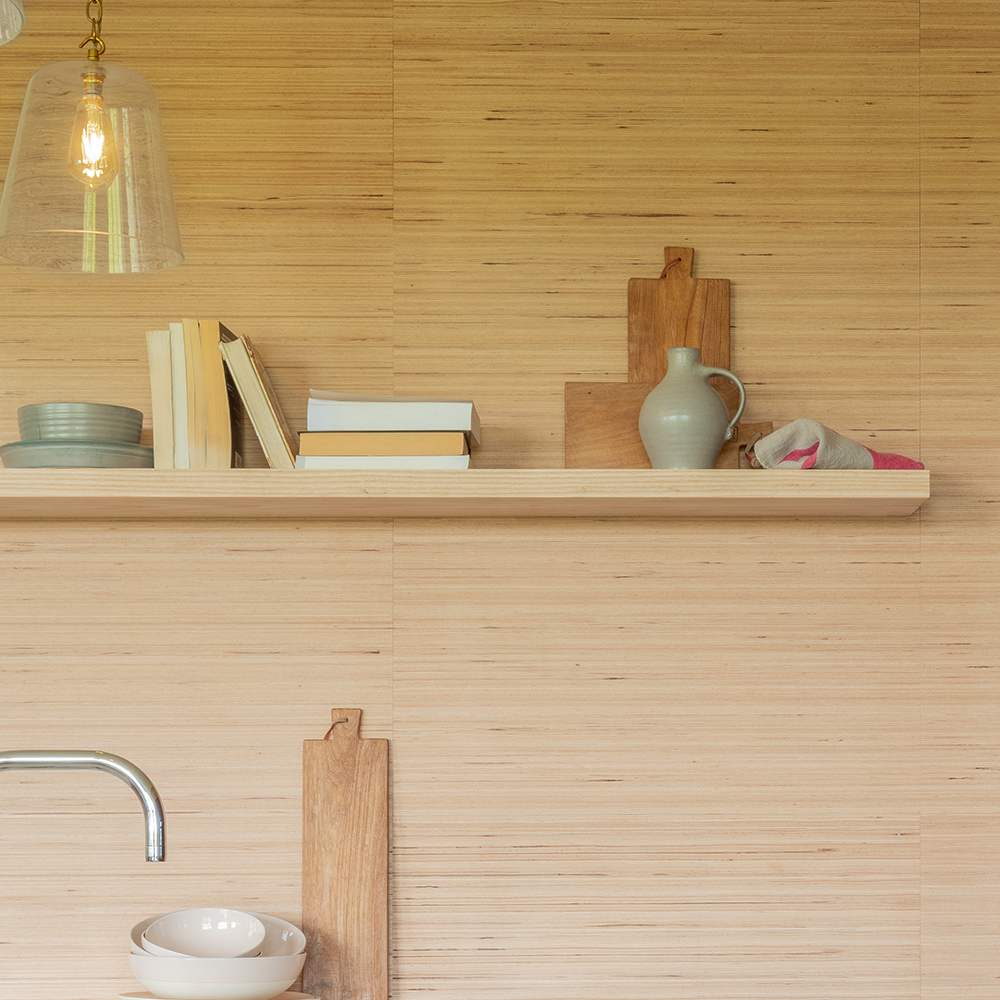 A wooden kitchen shelf holds stacked plates, books, a ceramic jug, and a folded towel. Wooden cutting boards stand upright behind the items. A glass pendant light hangs above.