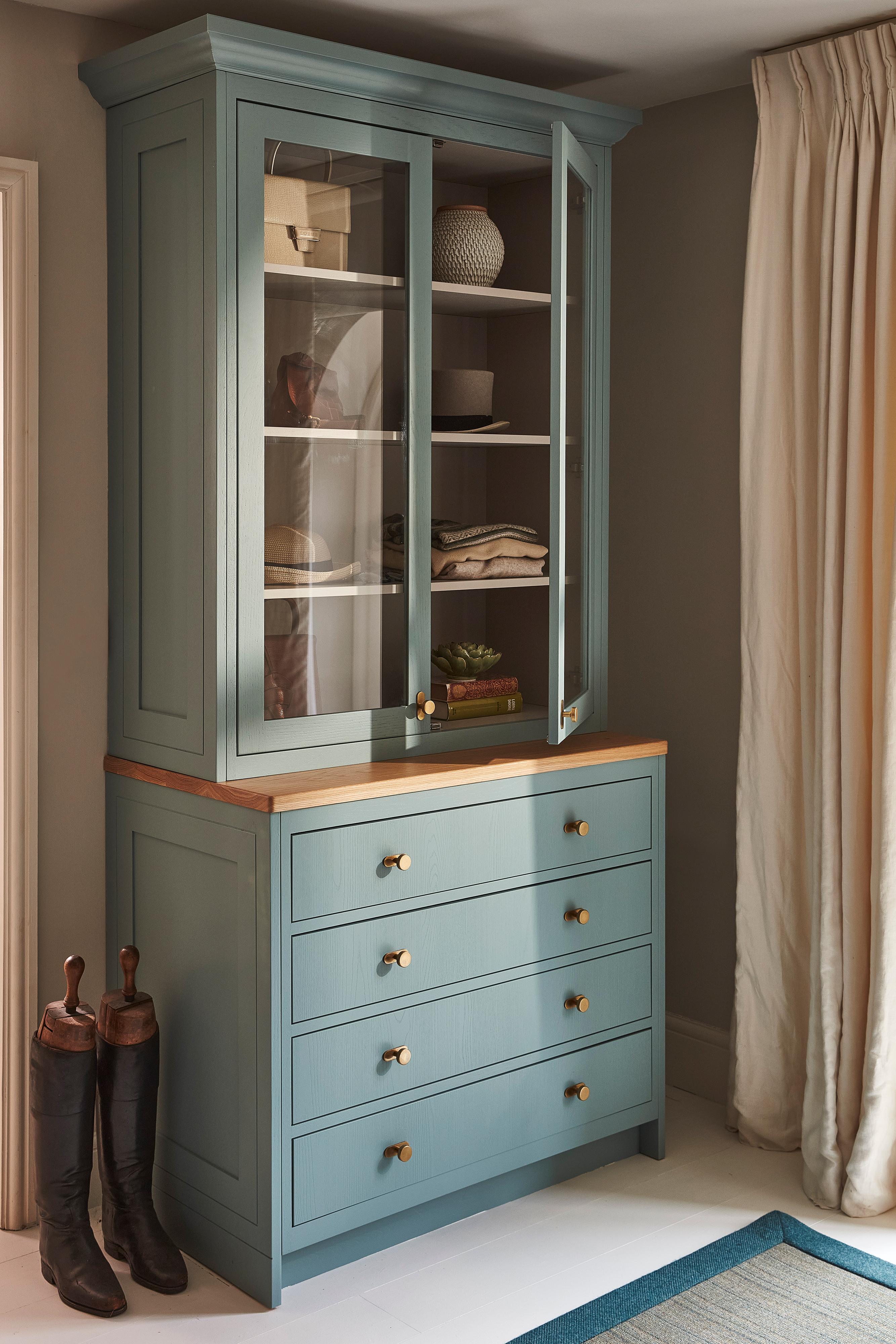 A tall blue cabinet with glass doors on top and four drawers below stands in a corner. Decorative items are displayed on the shelves. Brown boots are placed on the floor next to the cabinet. Cream curtains hang to the right.