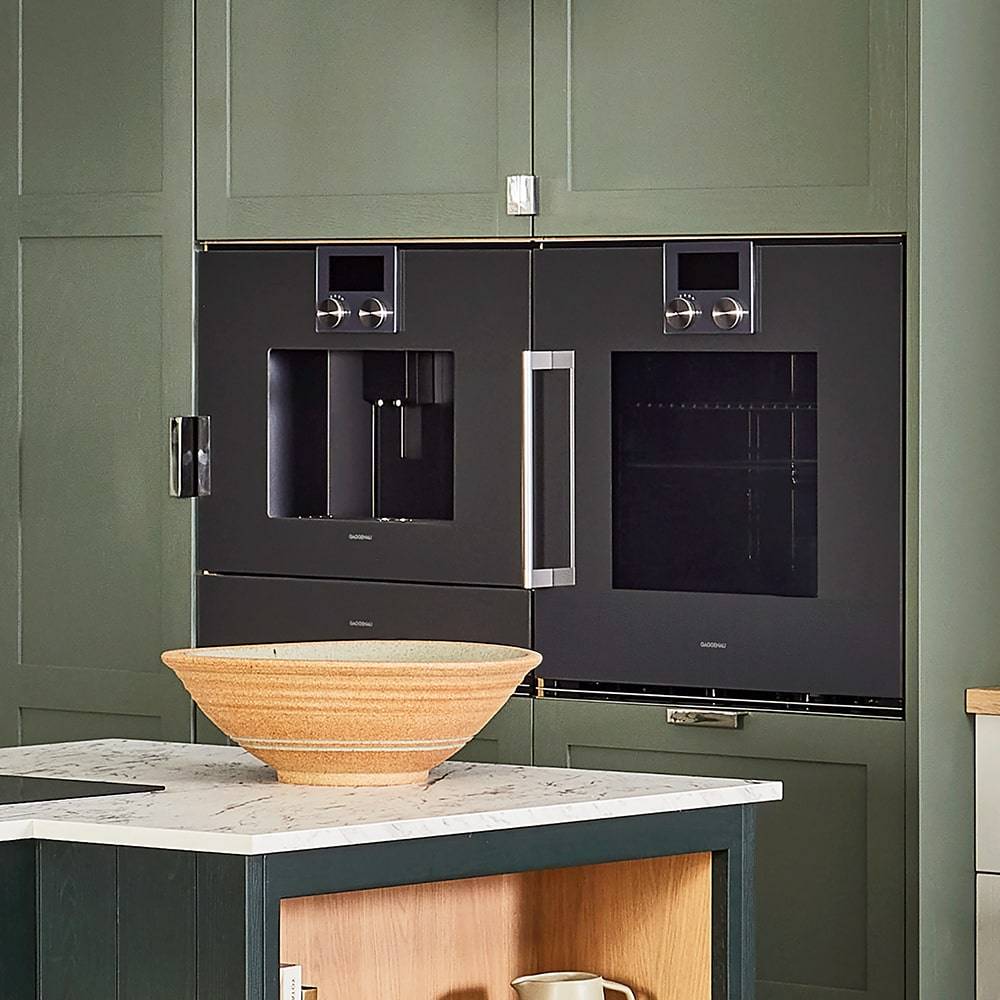 Built-in kitchen appliances with brushed metal finishes are integrated into matte green cabinets. A wooden bowl sits on a marble countertop of a kitchen island, contrasting with the dark appliance surfaces.