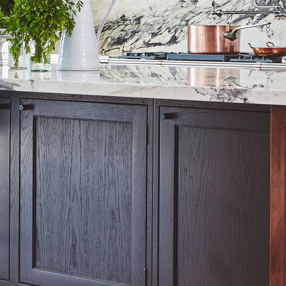  close-up of a modern kitchen features dark wood cabinets, a marble countertop, a copper pot on the stove, and a white pitcher with green foliage. The backsplash has a marbled pattern.