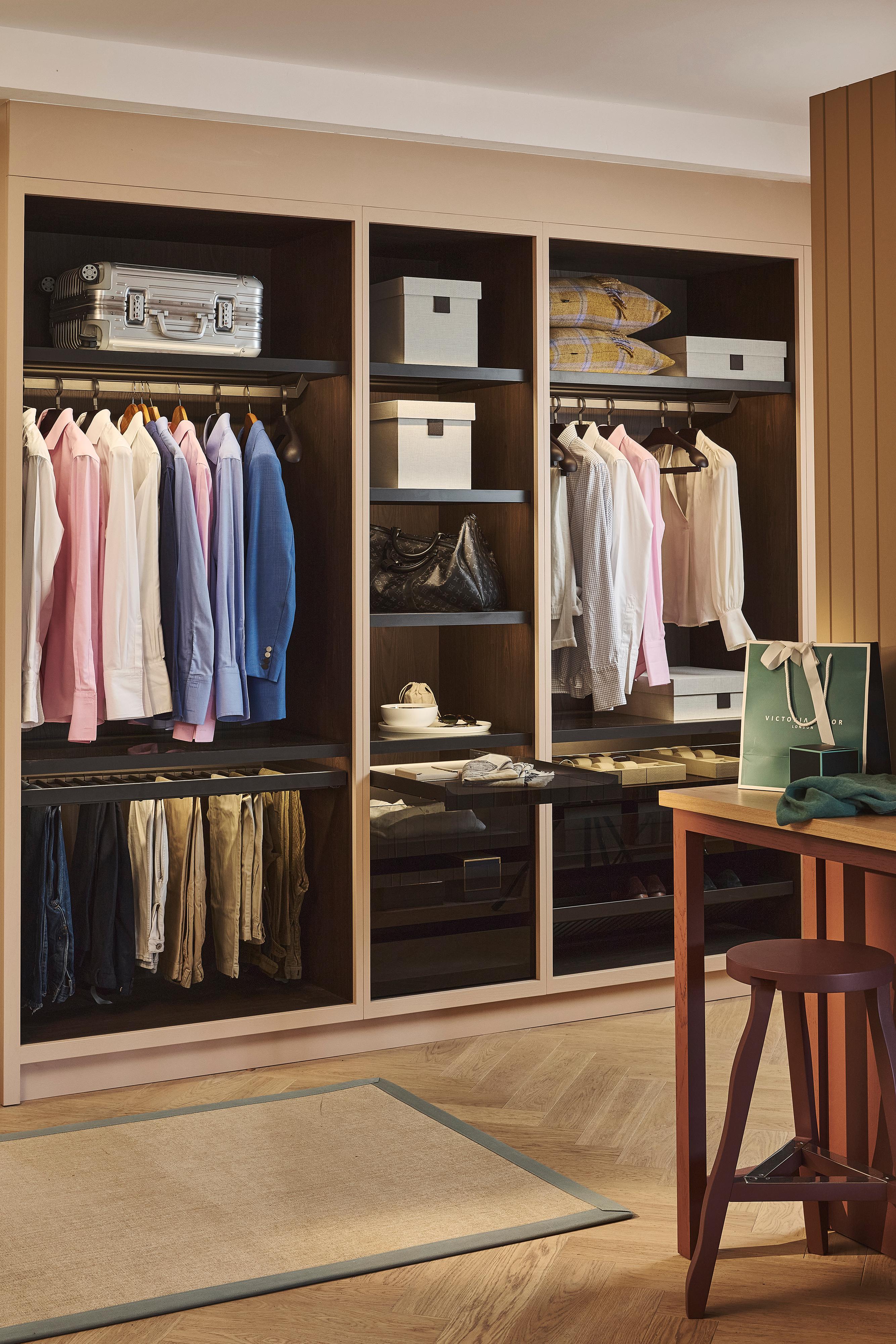 A neatly organized closet with shirts, jackets, and pants hanging, shelves with boxes and bags, folded clothes, and a suitcase. A wooden stool and shopping bag are visible in the foreground.