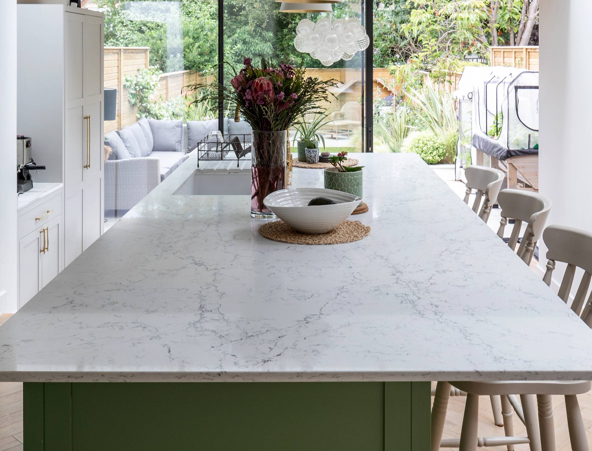 A modern kitchen with a large white marble island, light wood chairs, a vase of flowers, and a bowl. Glass doors in the background open to a patio with outdoor seating and a garden view.