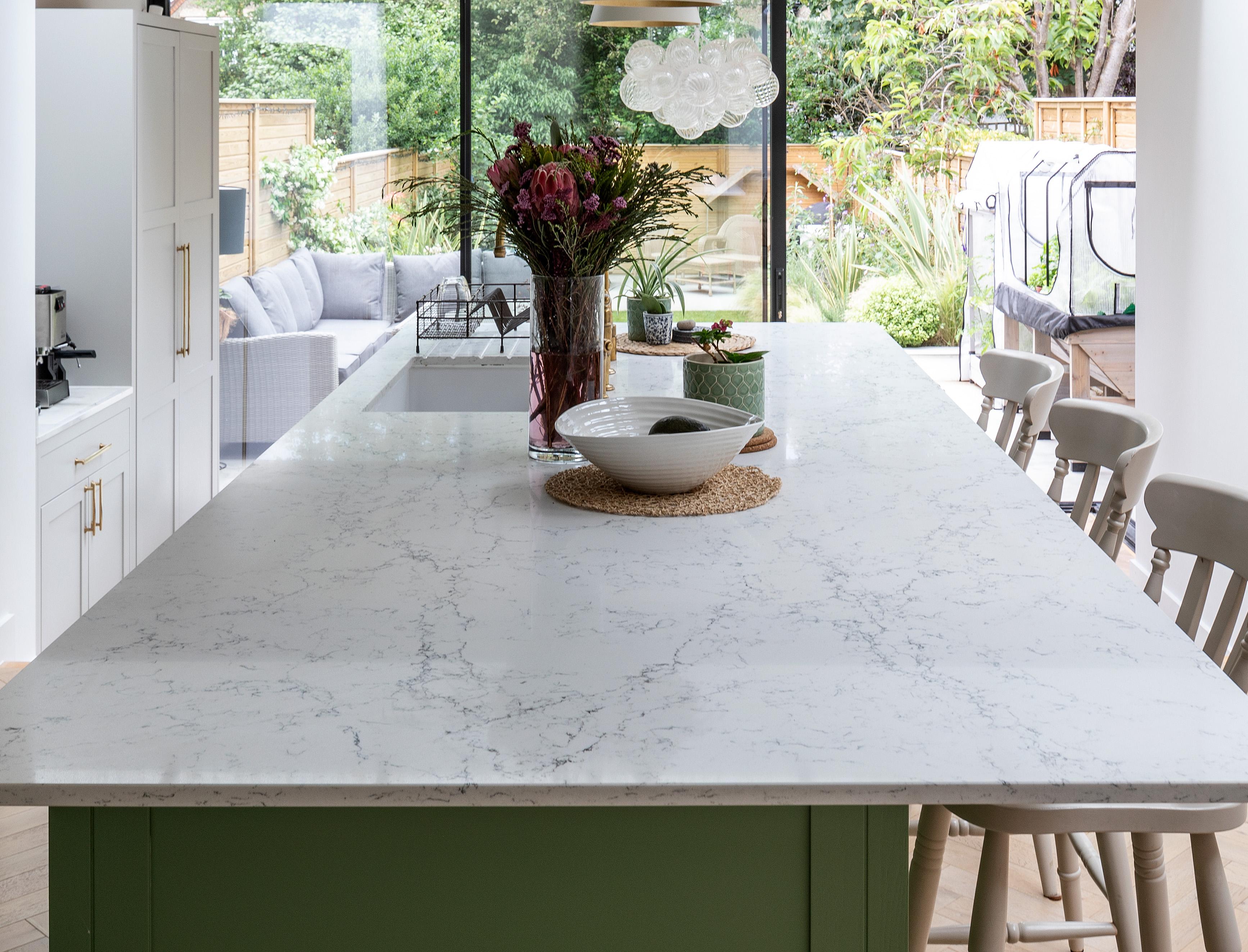 A modern kitchen with a large white marble island, light wood chairs, a vase of flowers, and a bowl. Glass doors in the background open to a patio with outdoor seating and a garden view.
