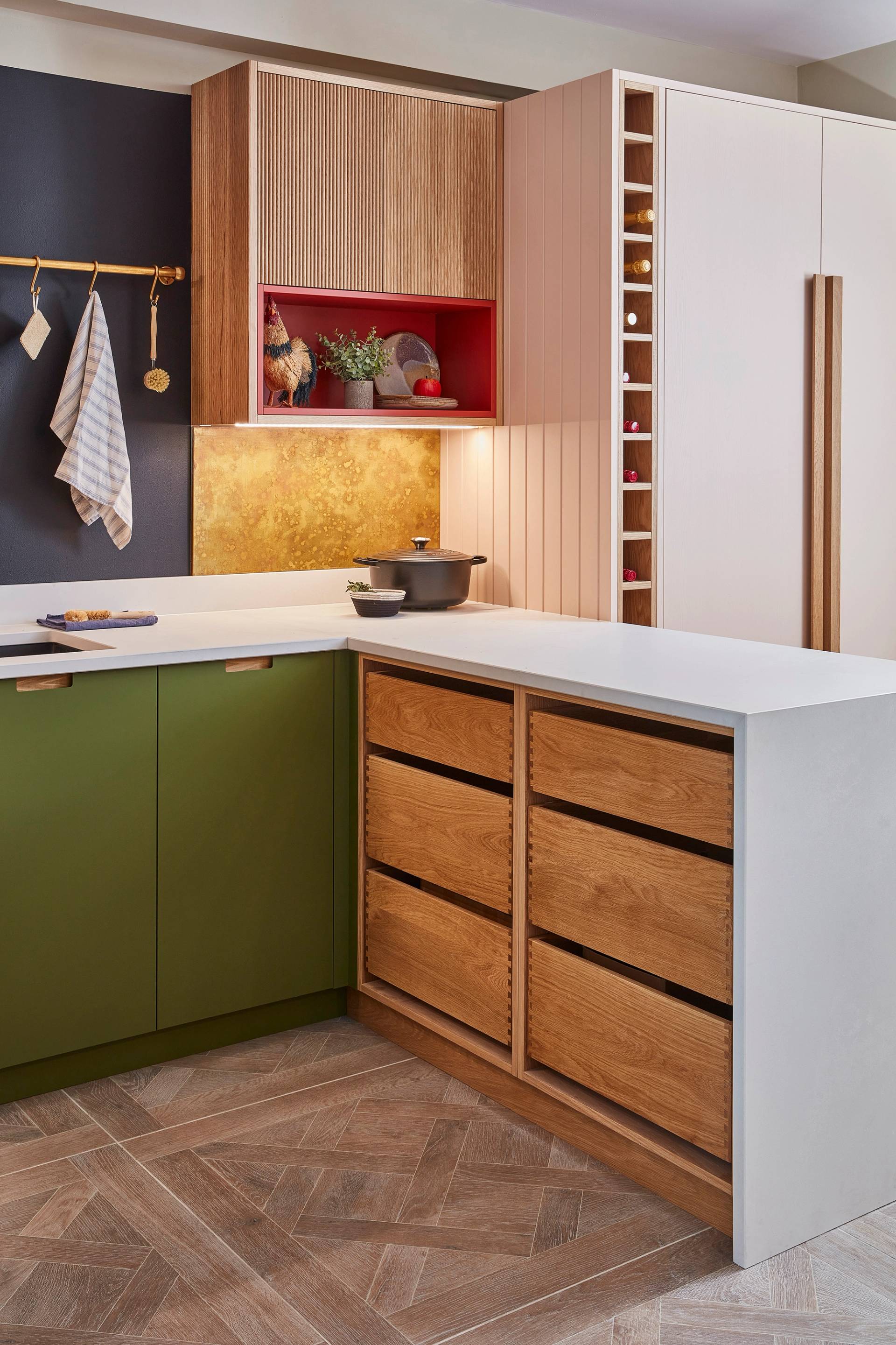 Modern kitchen with green cabinets, a blackboard wall listing groceries, and a gold faucet over a black sink. A wooden rod holds kitchen tools, herbs, and a striped towel. A bottle and sponge sit on the counter.