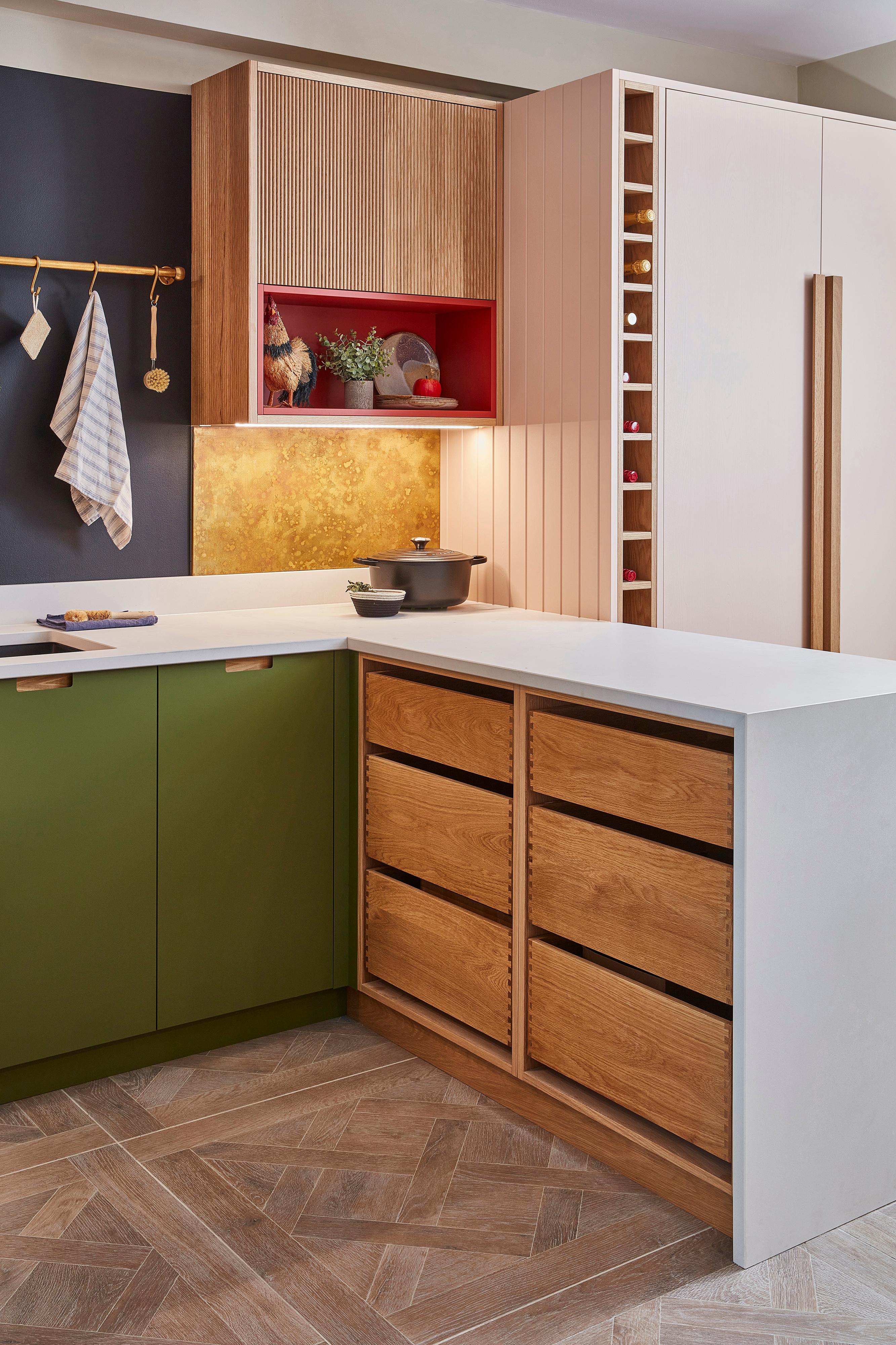 Modern kitchen with green cabinets, a blackboard wall listing groceries, and a gold faucet over a black sink. A wooden rod holds kitchen tools, herbs, and a striped towel. A bottle and sponge sit on the counter.