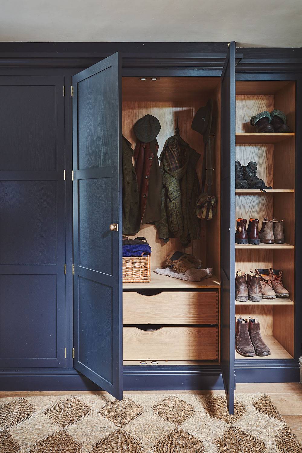 A stylish dark blue wardrobe is open, revealing hanging coats and a hat inside. To the right, there are shelves filled with shoes and boots. A woven basket sits on one of the two wooden drawers. The floor features a patterned rug.
